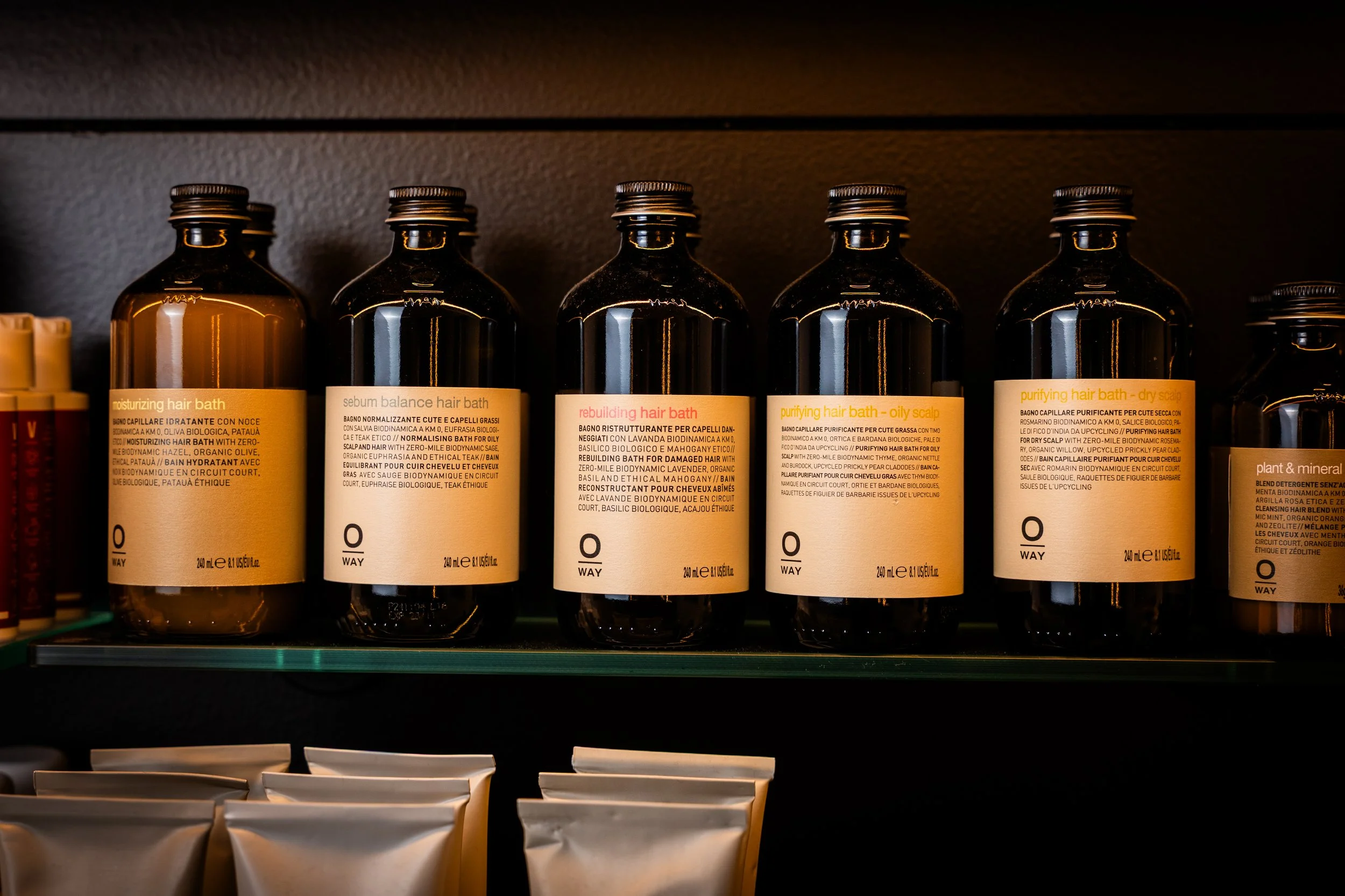Shelf with five dark brown bottles of hair bath products, each with a beige label describing different hair care functions, in a store setting.