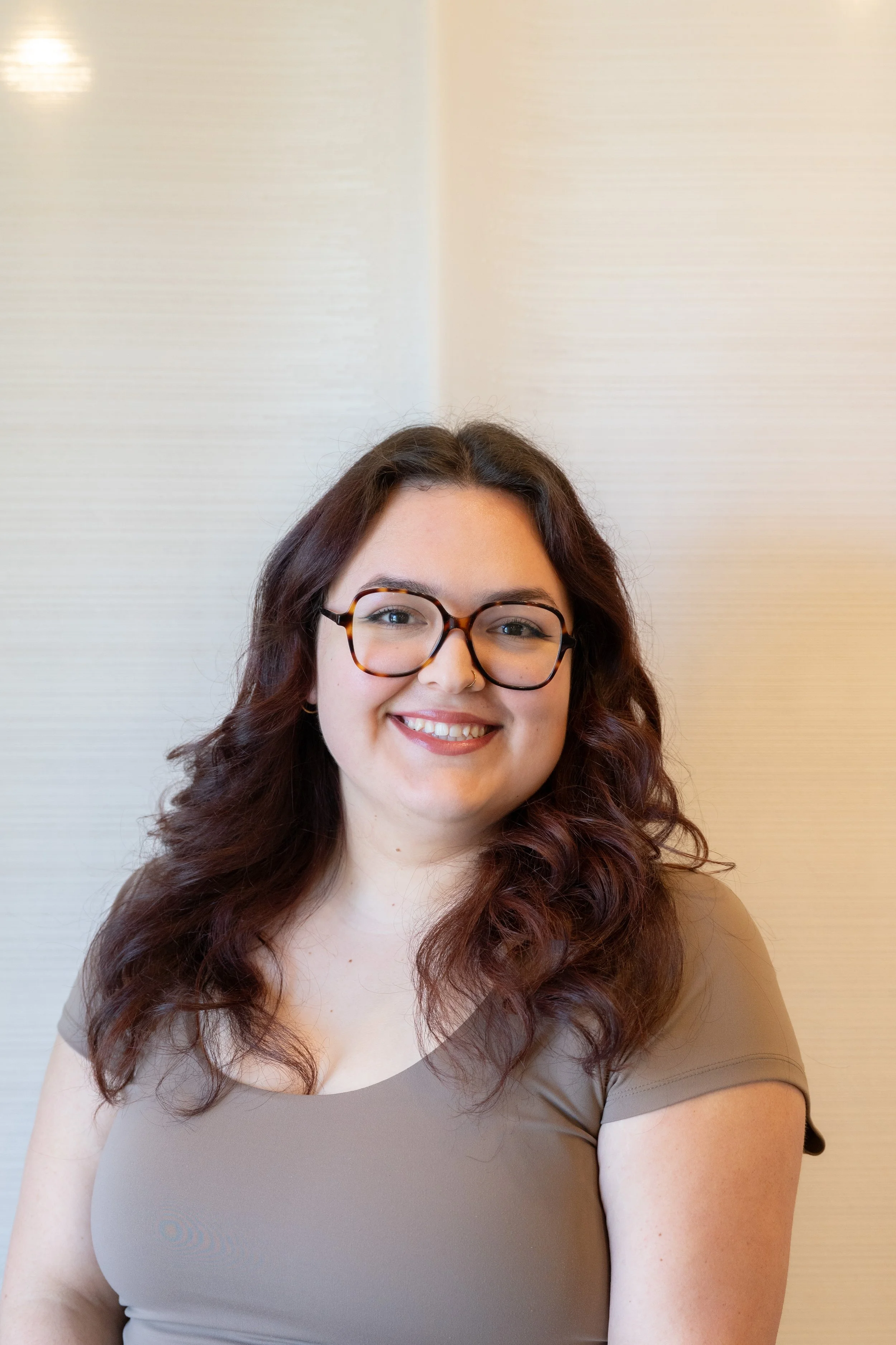A woman with long, wavy brown hair, wearing glasses and a tan short-sleeve top, smiling at the camera against a light-colored textured wall background.