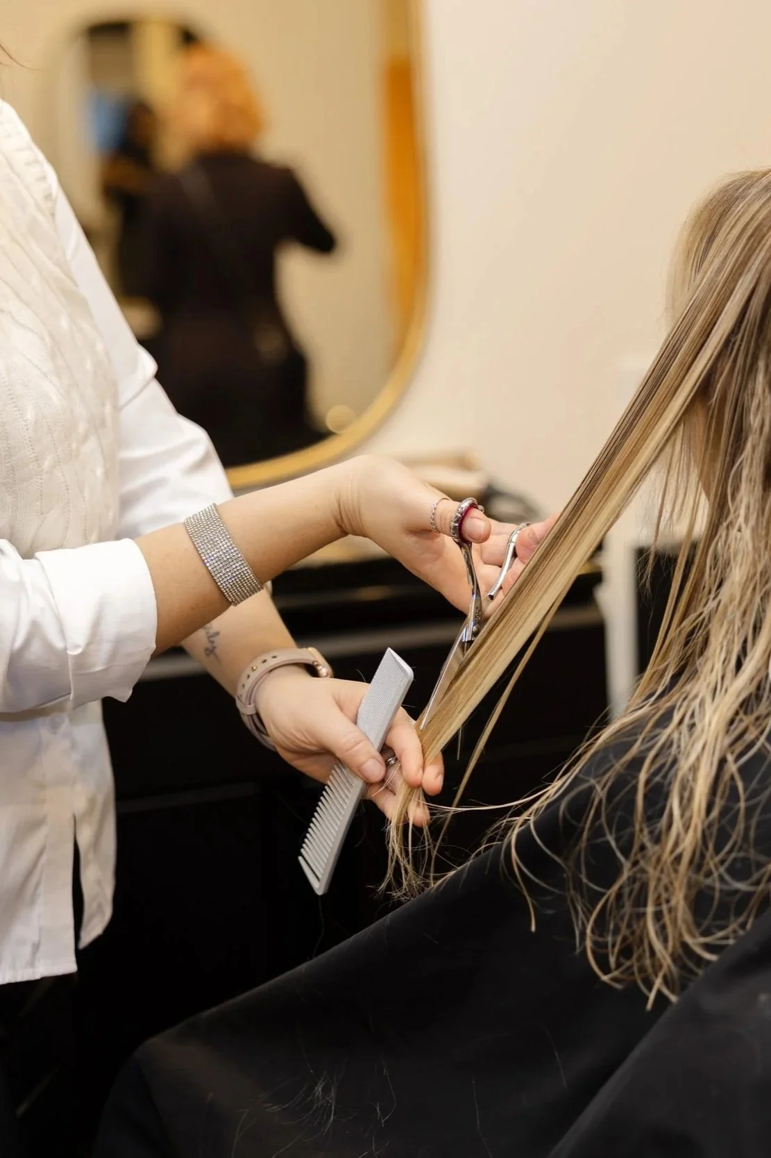 A hairstylist cutting a client's long blonde hair with scissors and holding the hair with a comb in a salon. The background shows a mirror with a blurred reflection of another person.