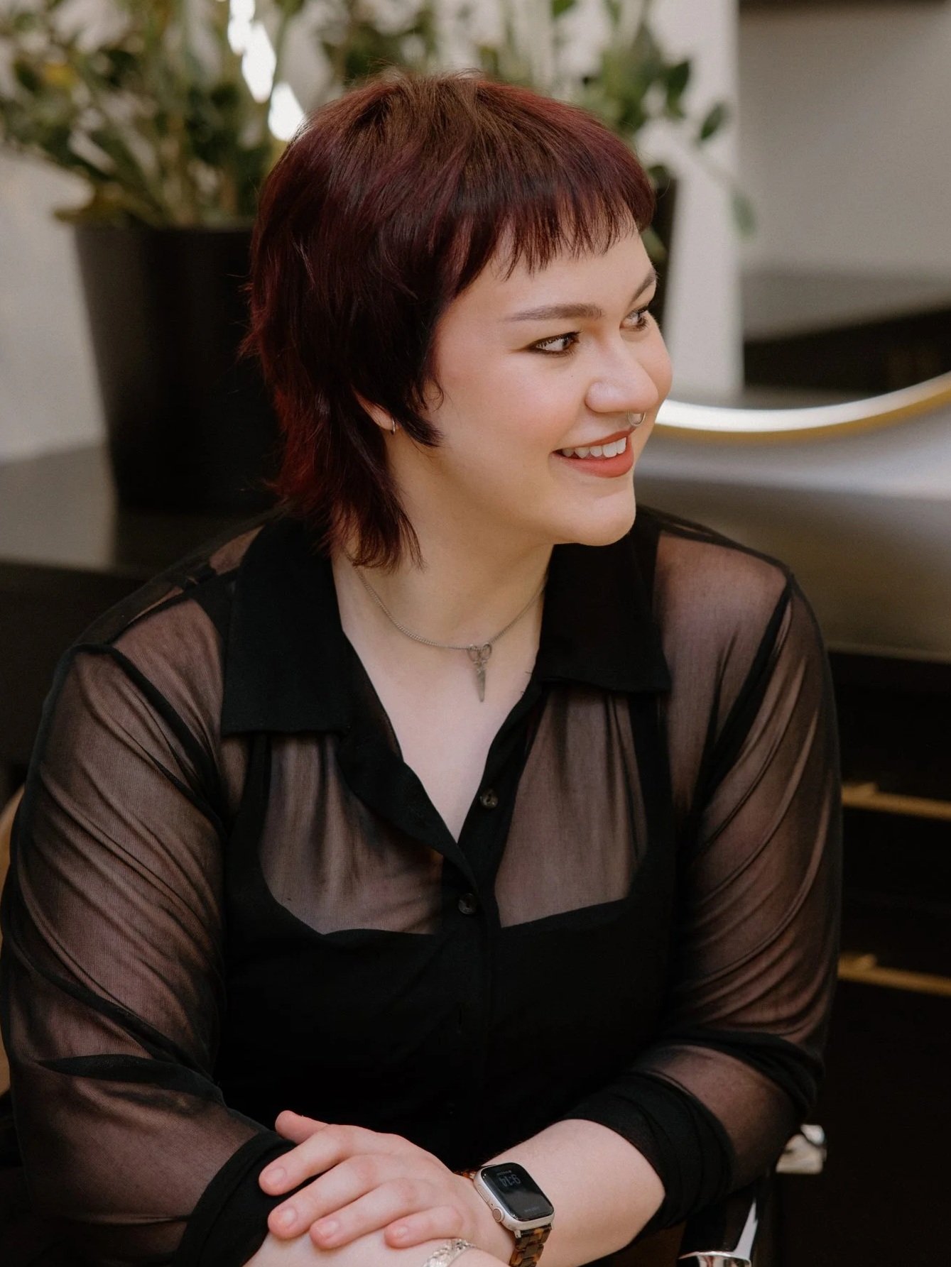 person with short red hair and light makeup, wearing earrings, a necklace, and a black top, smiling at the camera in an indoor setting.