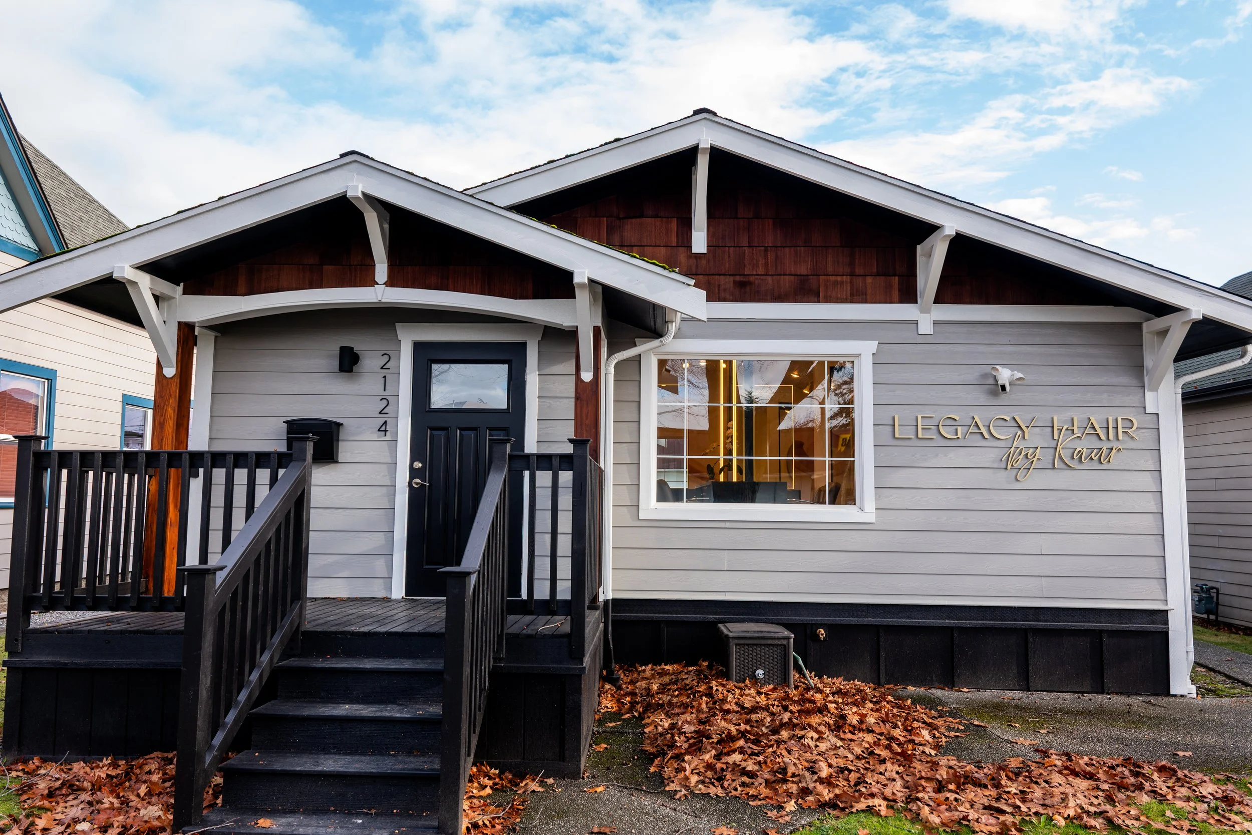 Exterior view of a hair salon named 'Legacy Hair by Kaur' with a black front door, large window, and a small porch with black stairs. The building has gray siding with wooden accents and the salon's name displayed on the wall. There are fallen leaves on the ground and a partly cloudy sky above.