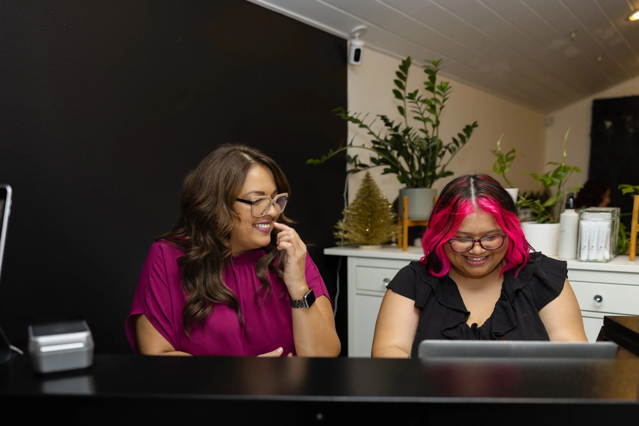 Two women sitting at a reception desk, smiling and looking at a tablet.