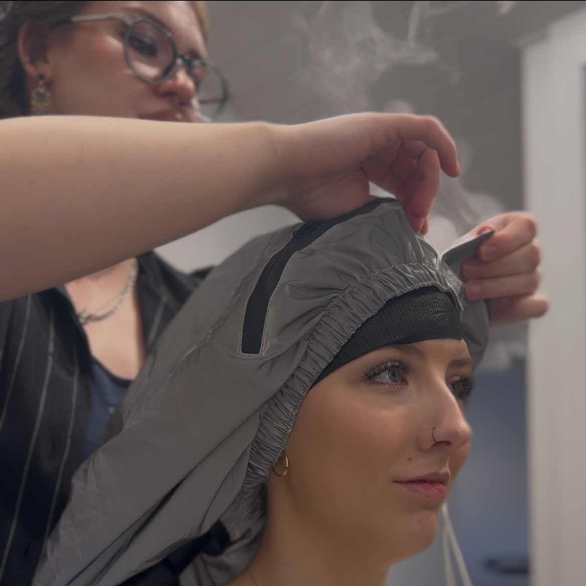 A woman getting her hair colored or styled by a stylist in a salon, with a black cap and a gray cape around her shoulders.