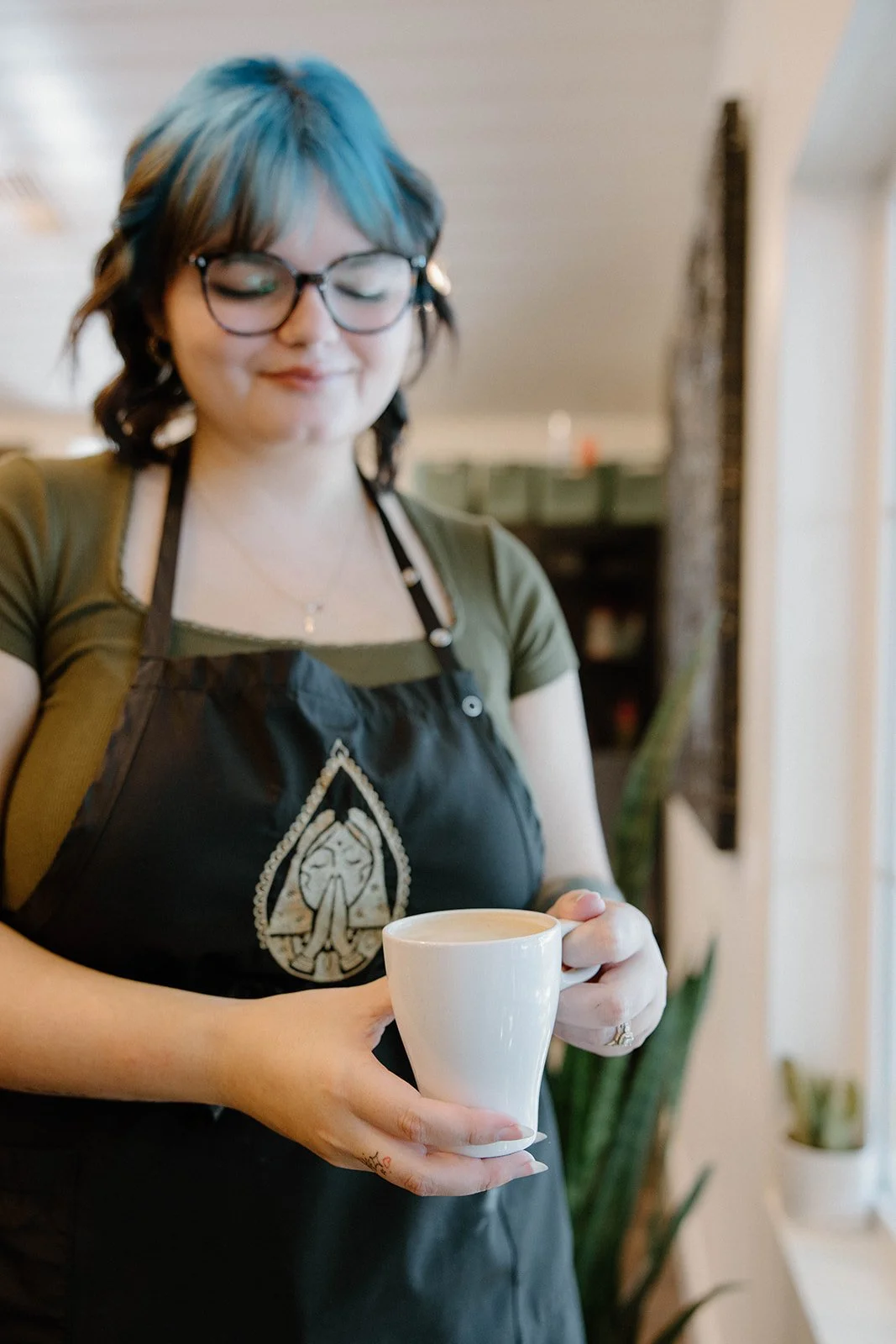 A woman with glasses and colorful blue and brown hair in a coffee shop, wearing a green shirt and a black apron with a logo, holding a white mug of coffee or tea.