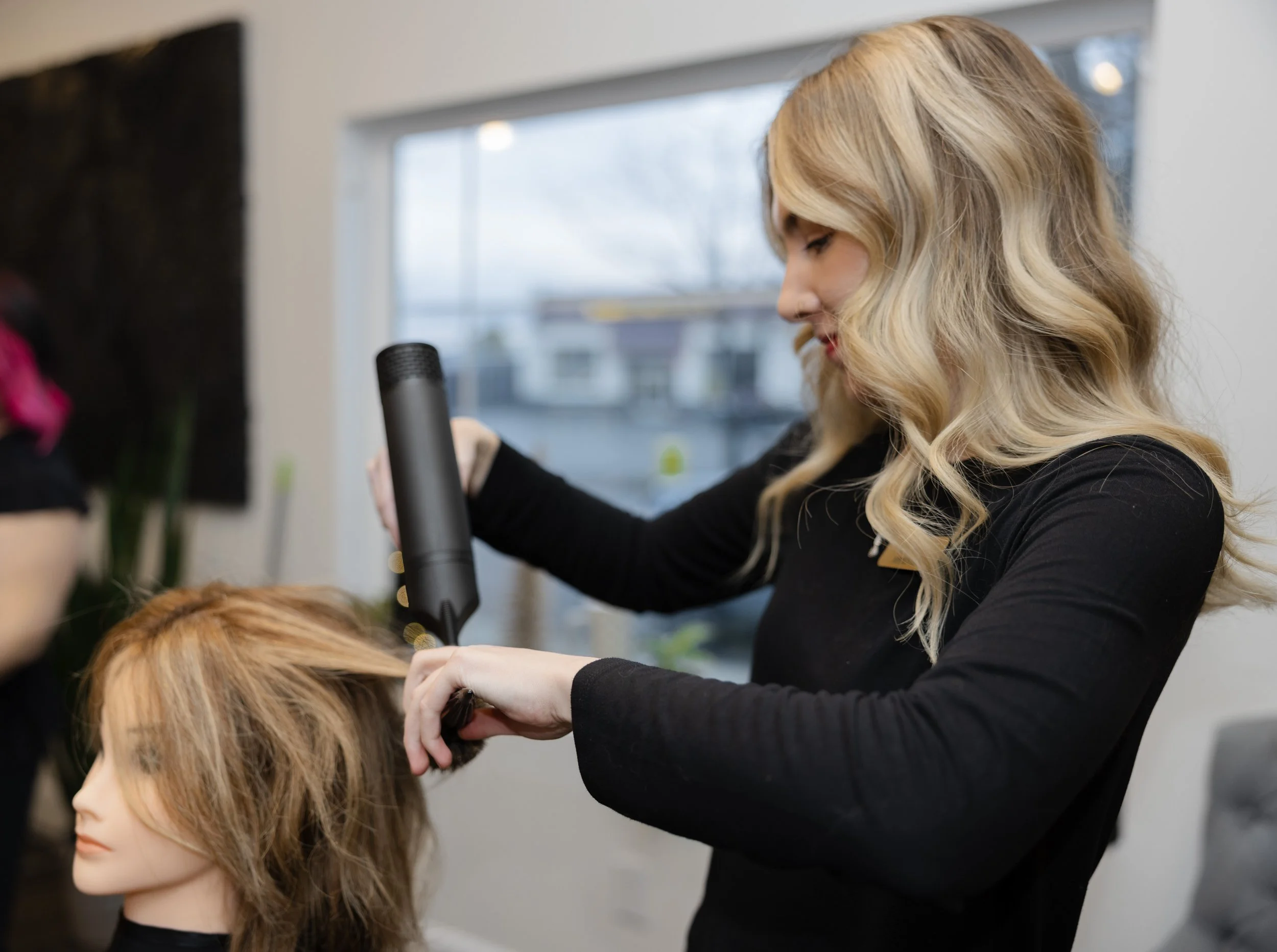 Woman with blonde hair styling a mannequin with brown hair in a salon.