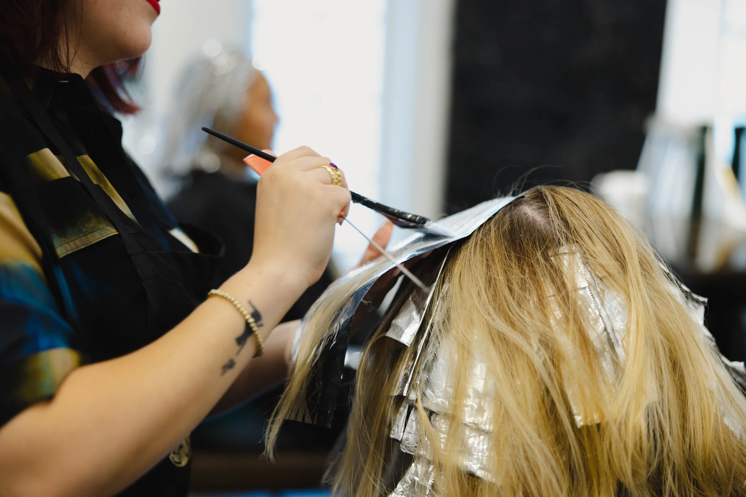 A woman getting hair color or highlights applied by a stylist in a salon, with hair foils and aluminum wraps in her blonde hair.