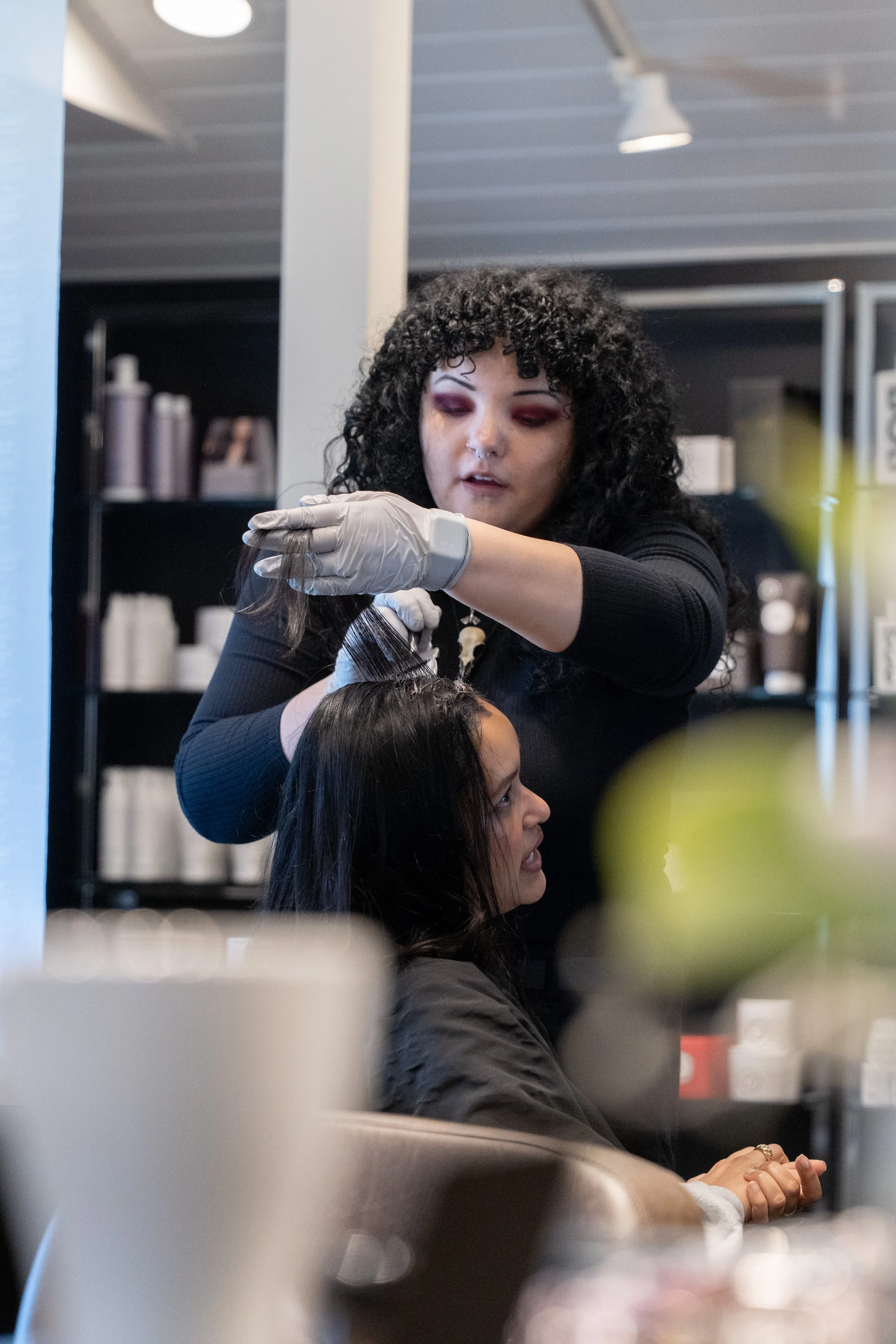 Hair stylist with curly black hair and purple eye makeup dyeing a woman's long dark hair inside a salon.