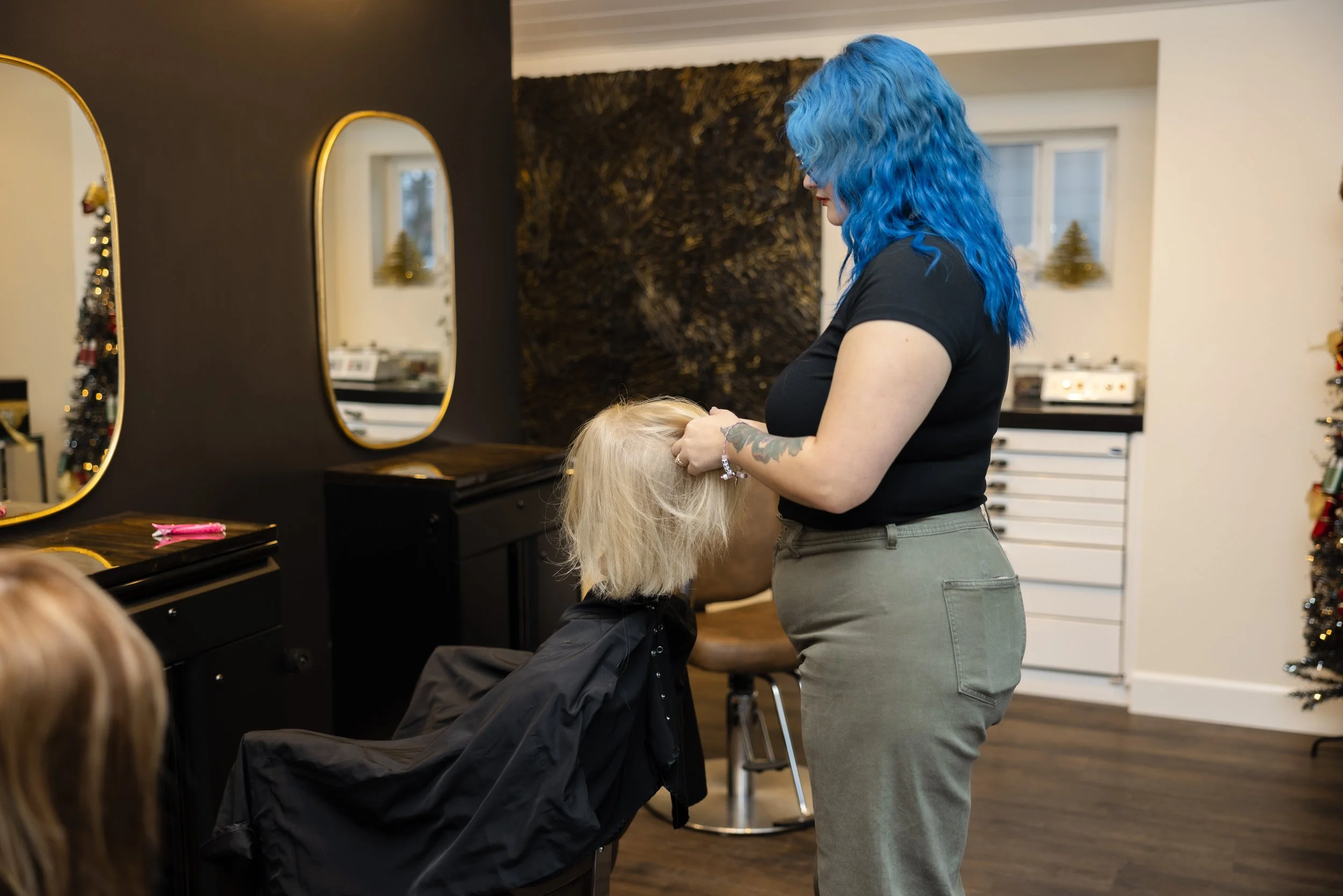 A woman with vibrant blue hair standing in a salon, styling or inspecting the blonde hair of a seated client, with the salon decorated for Christmas in the background.