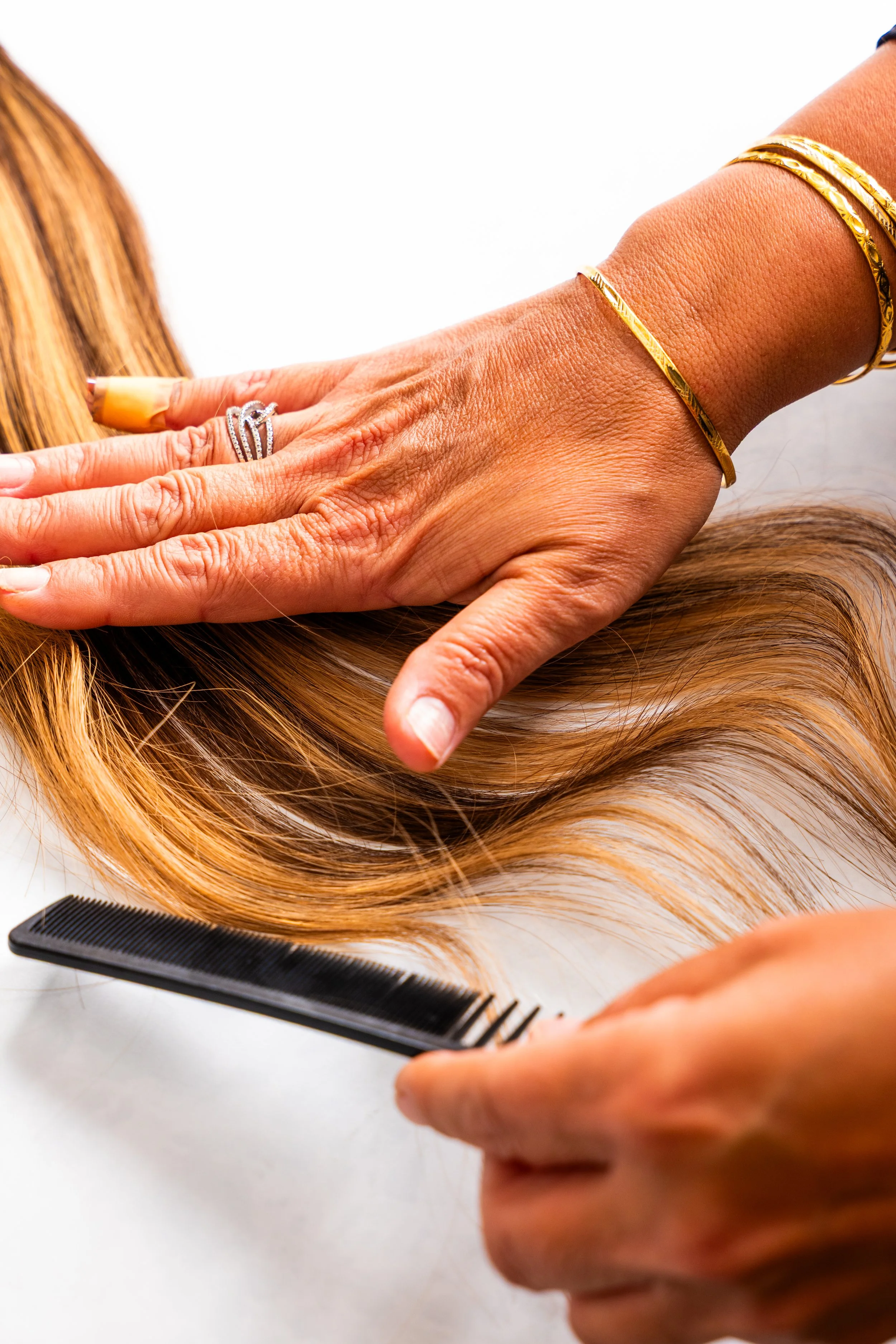 Close-up of a person's hand with gold jewelry and rings, gently brushing through and styling long, blonde hair, with a black hair comb nearby on a white surface.