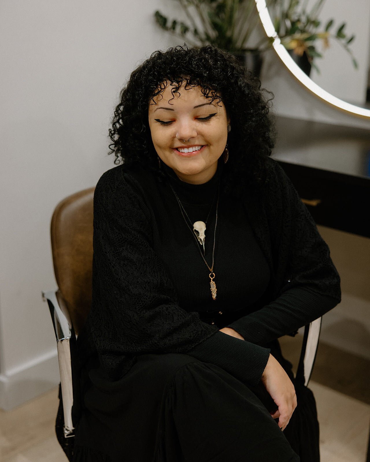 A woman with curly black hair and makeup, holding scissors toward the camera with a confident smile, wearing a black top with lace sleeves and a belt.
