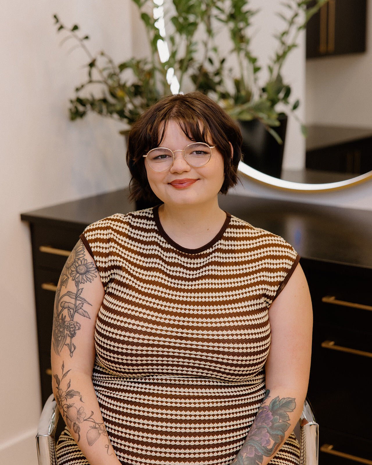 A young woman with curly blue hair, wearing glasses, a black t-shirt, and khaki pants, sitting on a leather chair in a salon or studio with mirrors and a black and gold decor.