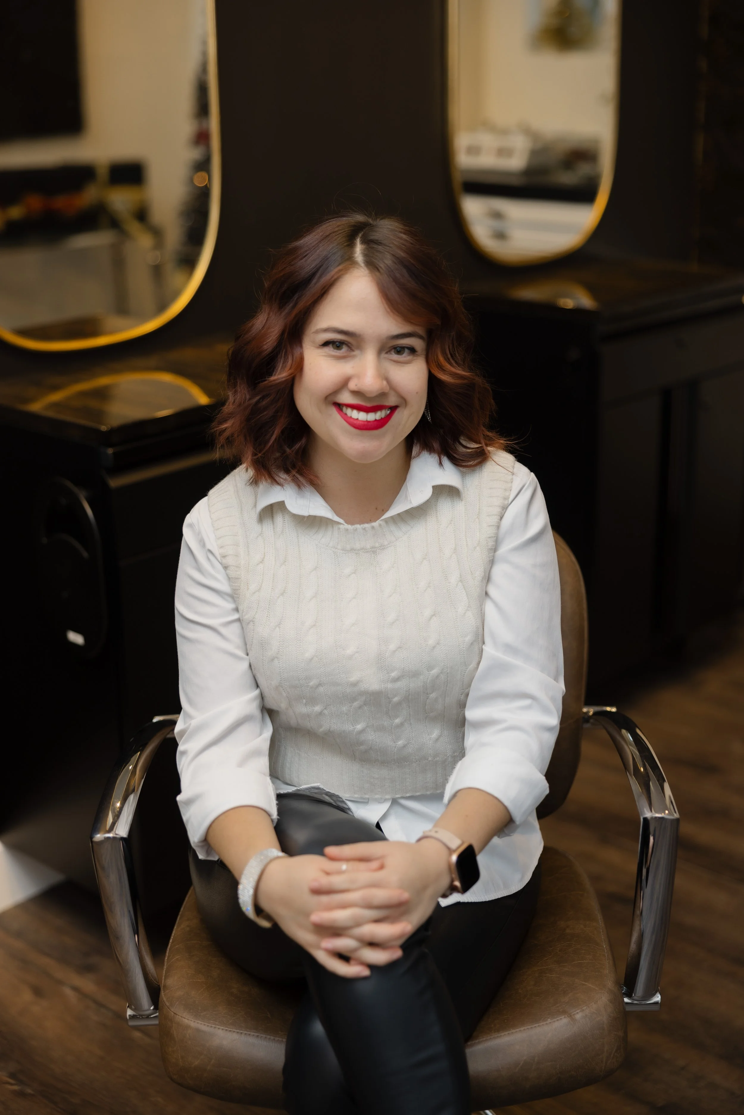 A woman with red hair and bright red lipstick sitting on a salon chair, smiling, in front of black mirrors with gold trim, in a hair salon.