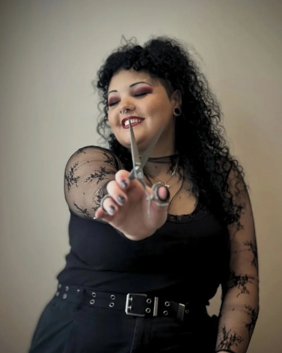 A woman with curly black hair and makeup, holding scissors toward the camera with a confident smile, wearing a black top with lace sleeves and a belt.