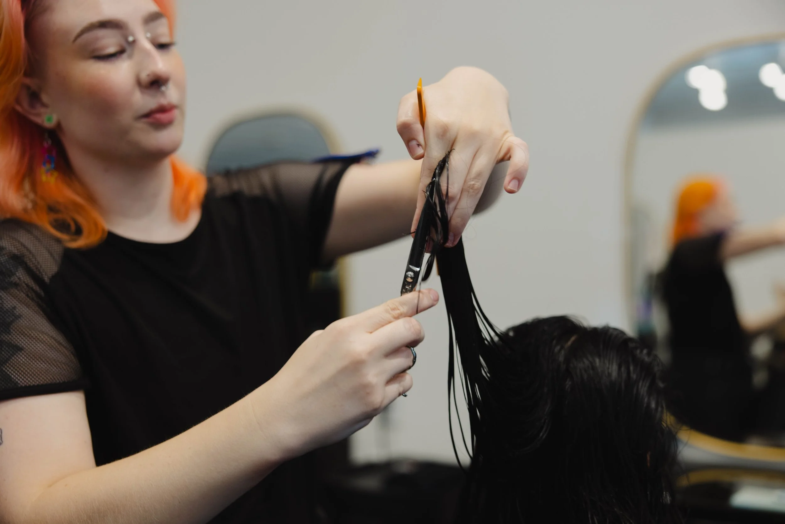 A hairstylist with orange hair, wearing a black top with mesh sleeves, trims a client's black, wet hair with scissors in a salon, with mirrors and salon equipment reflected in the background.