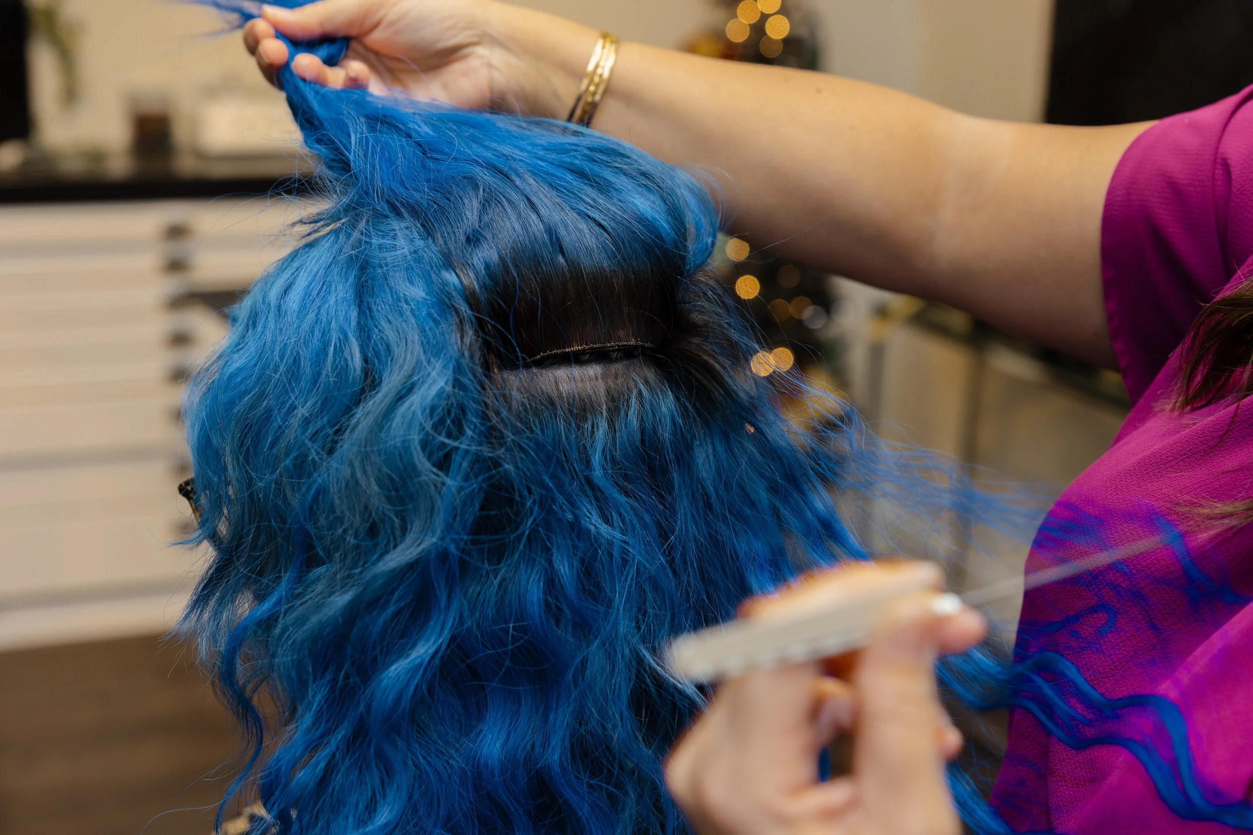 Person in pink shirt styling vibrant blue curly wig with a comb in a room with white cabinets and warm lighting.