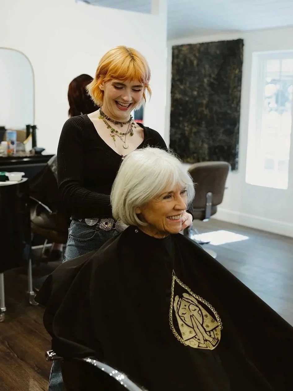 A hairstylist cutting a woman's gray hair in a salon, both smiling and looking happy.