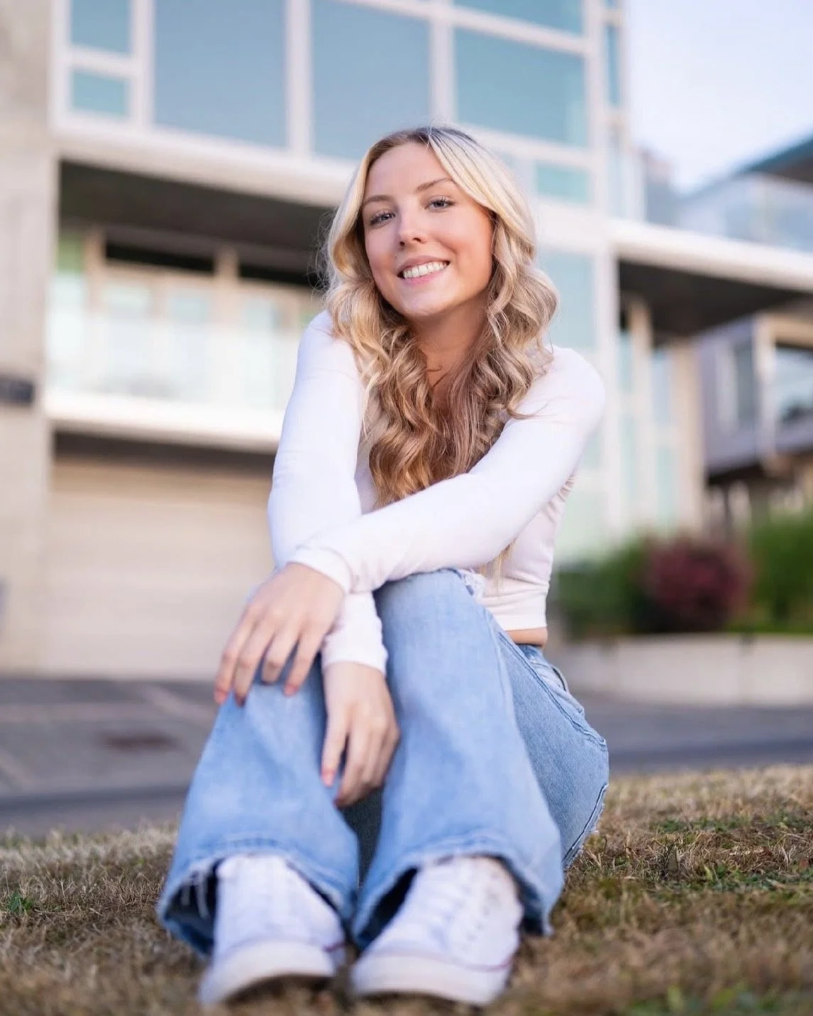 A young woman with long blonde hair, smiling, sitting on the grass in front of a modern apartment building