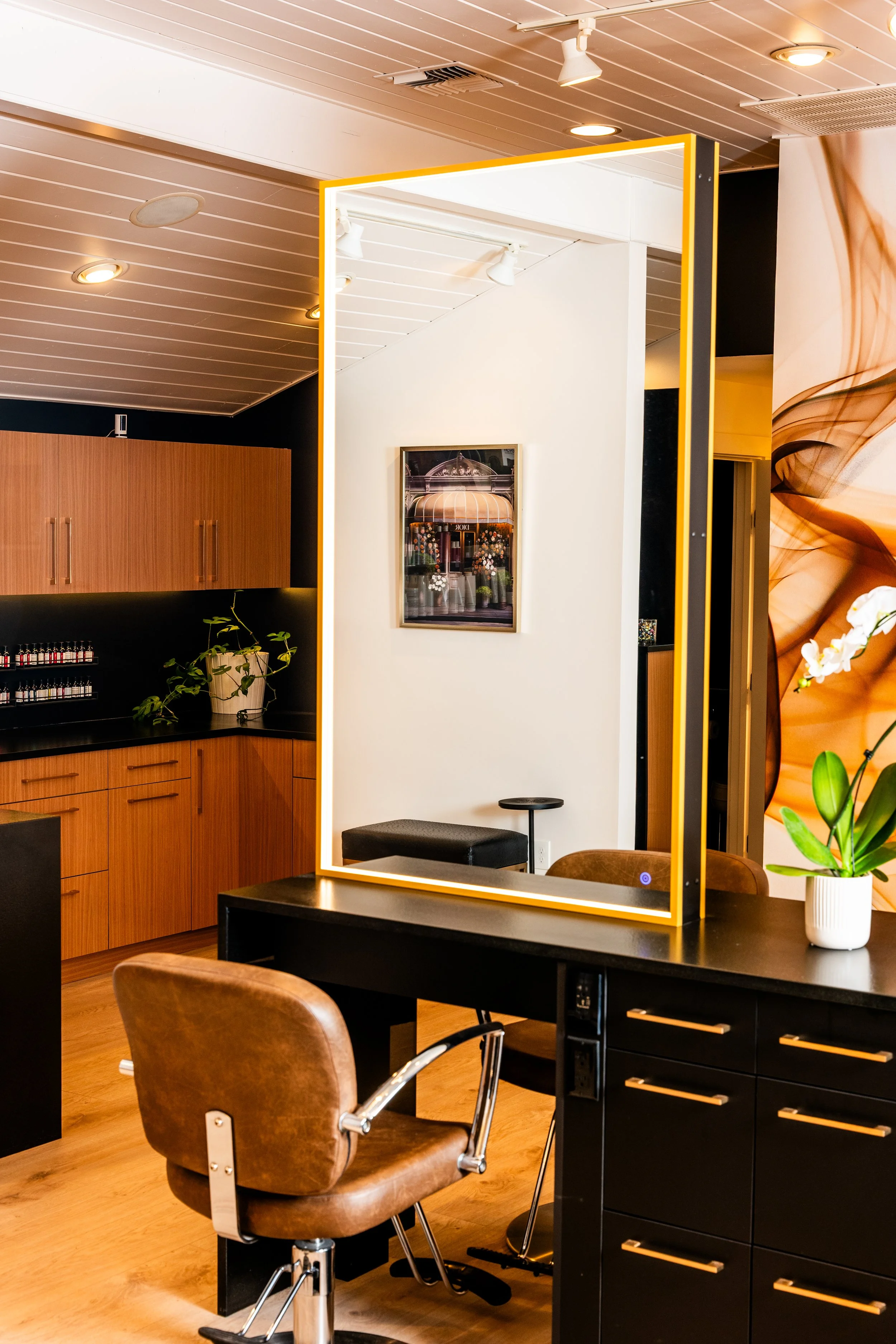 Interior of a hair salon with a black styling station, a brown leather swivel chair, a large illuminated mirror, and a white orchid plant on the counter.