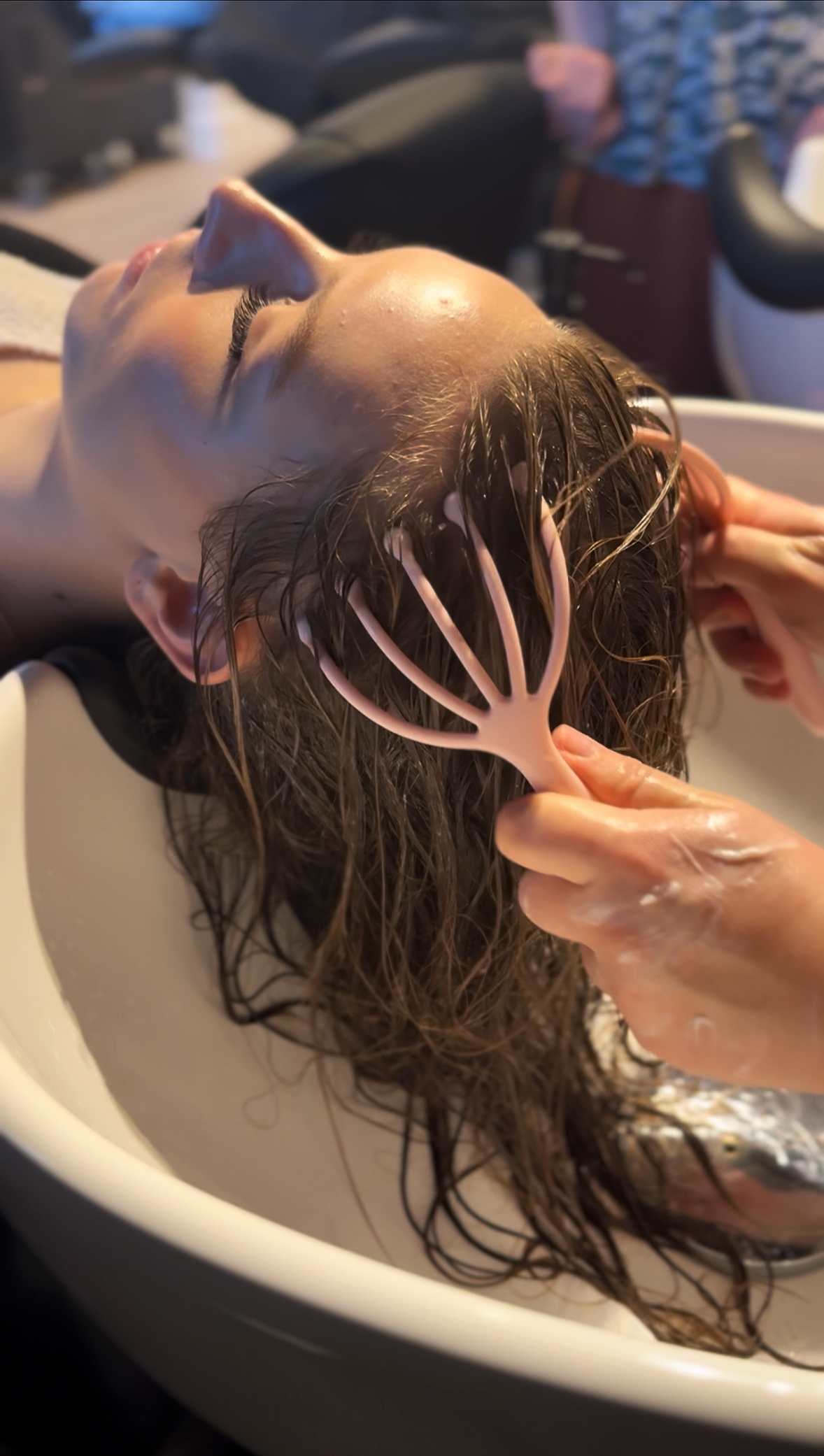 A woman with wet hair lying in a salon sink while a stylist shampoos her hair with a pink hair comb.