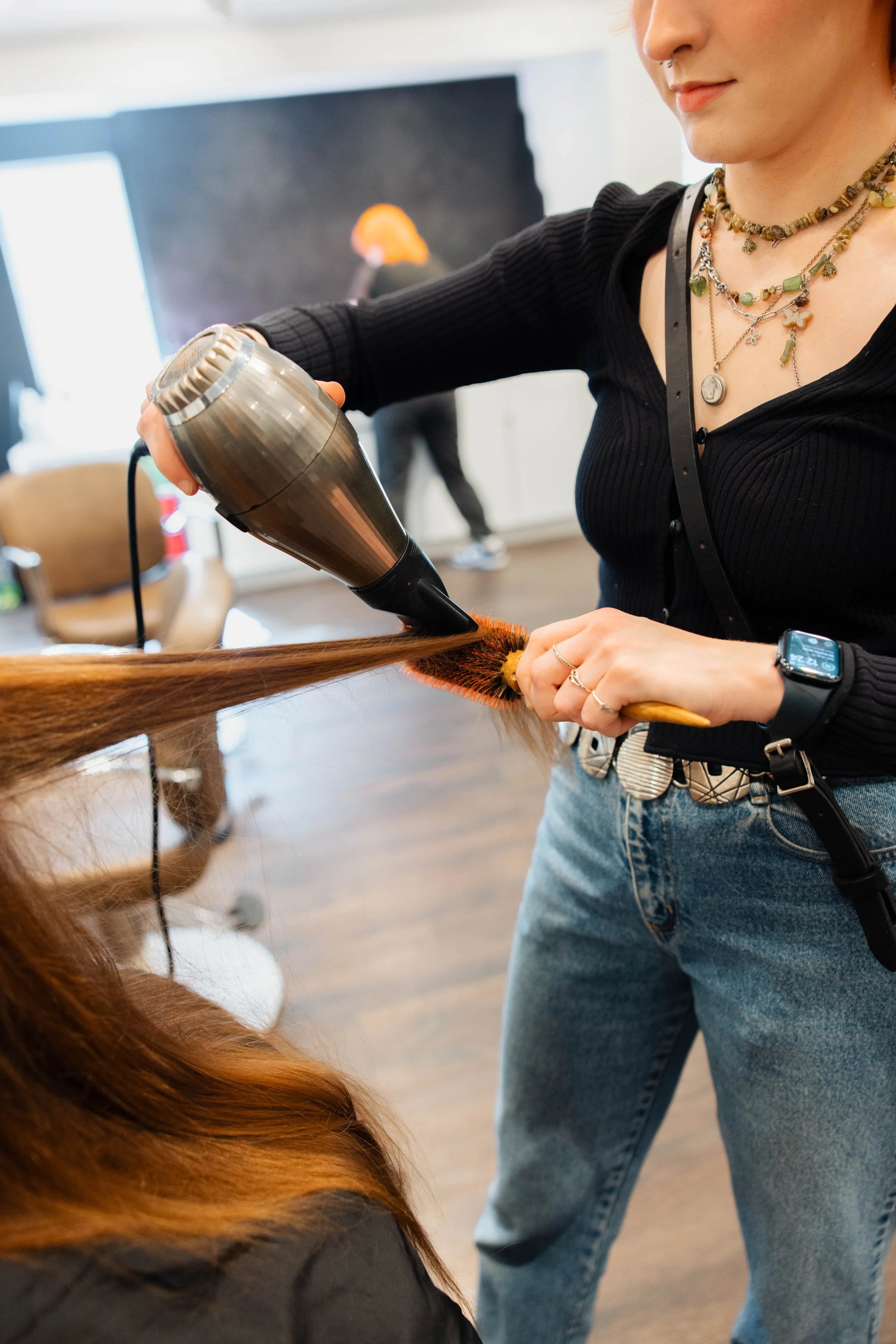 A hairdresser blow-drying a client's long, straight, reddish hair with a black and silver hairdryer at a salon.