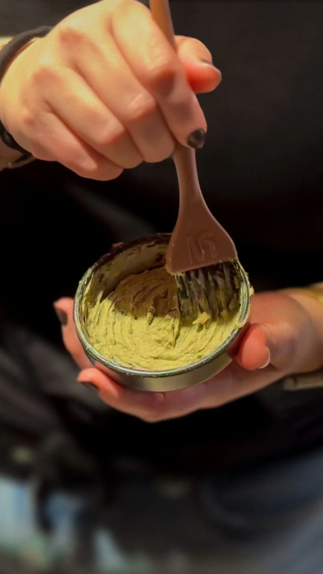 A person is holding a small jar of yellowish paste and using a silicone spatula to stir or scoop it.