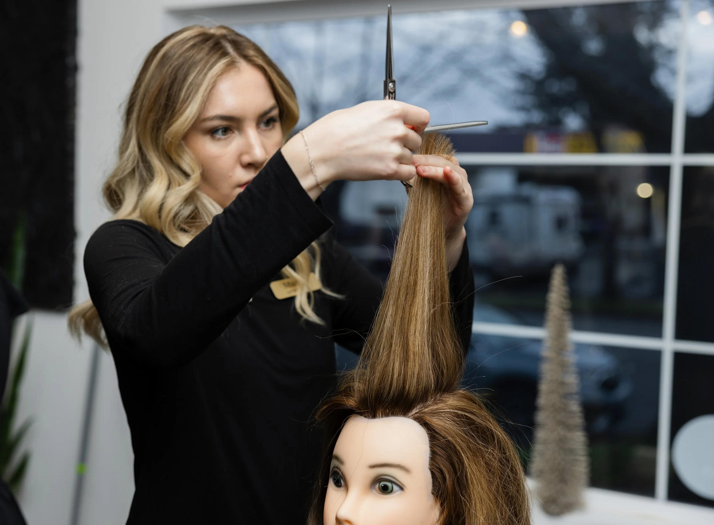 A hairstylist practicing haircutting skills on a mannequin head with long, brown hair inside a salon.