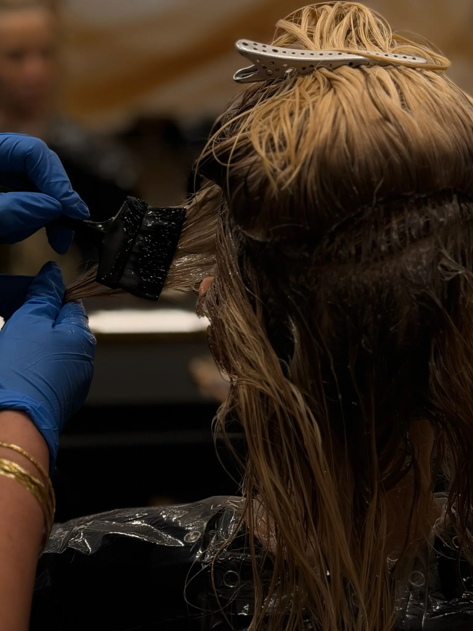 Hair stylist with curly black hair and purple eye makeup dyeing a woman's long dark hair inside a salon.