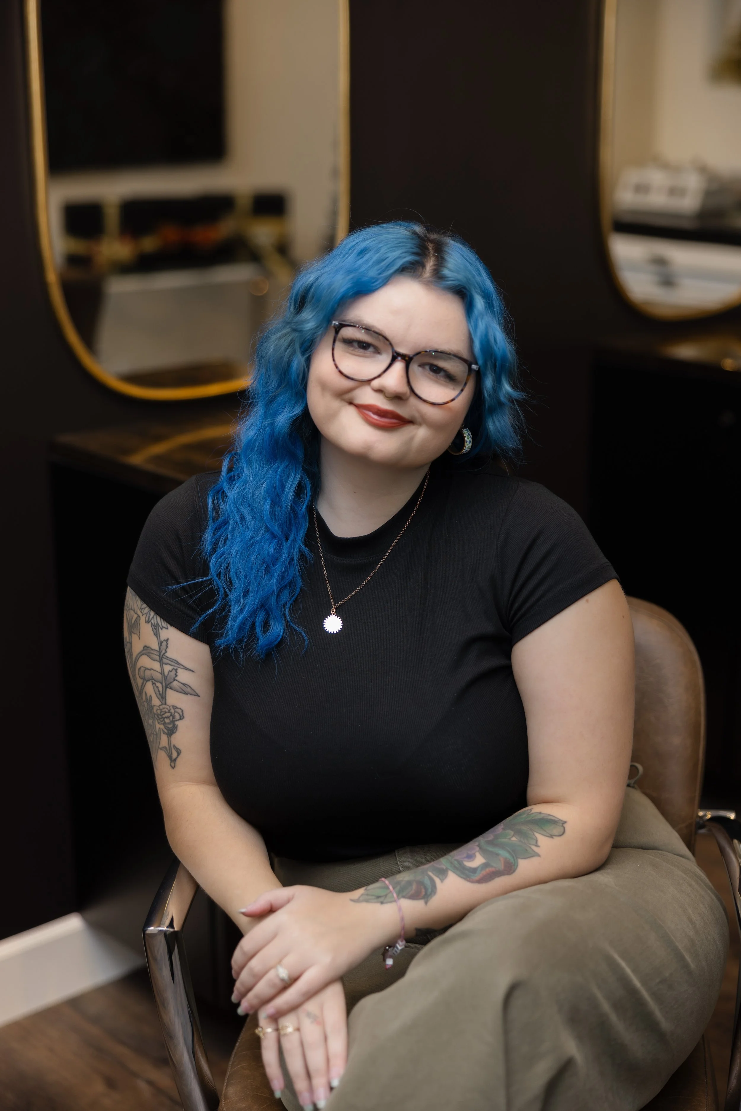 A young woman with curly blue hair, wearing glasses, a black t-shirt, and khaki pants, sitting on a leather chair in a salon or studio with mirrors and a black and gold decor.