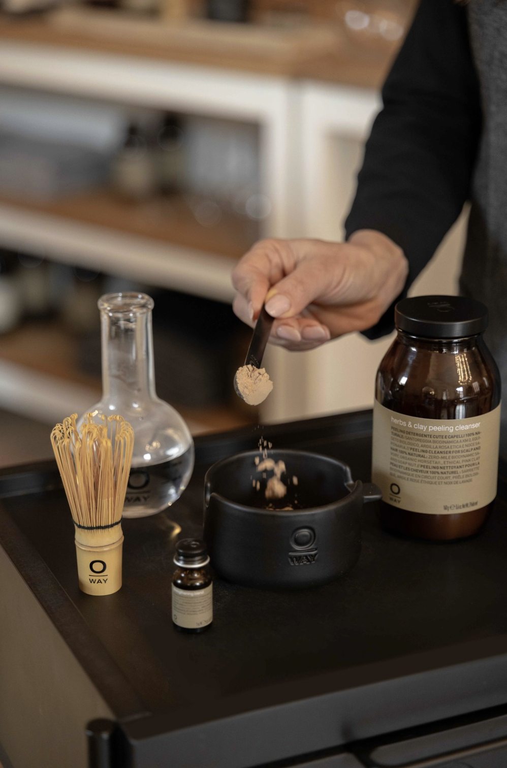 Person dispensing powder from a small container into a black bowl on a black tray, surrounded by a brown herbal and clay peeling cleanser jar, clear glass flask with liquid, a small brown bottle, and a bamboo whisk, in a skincare or spa setting.
