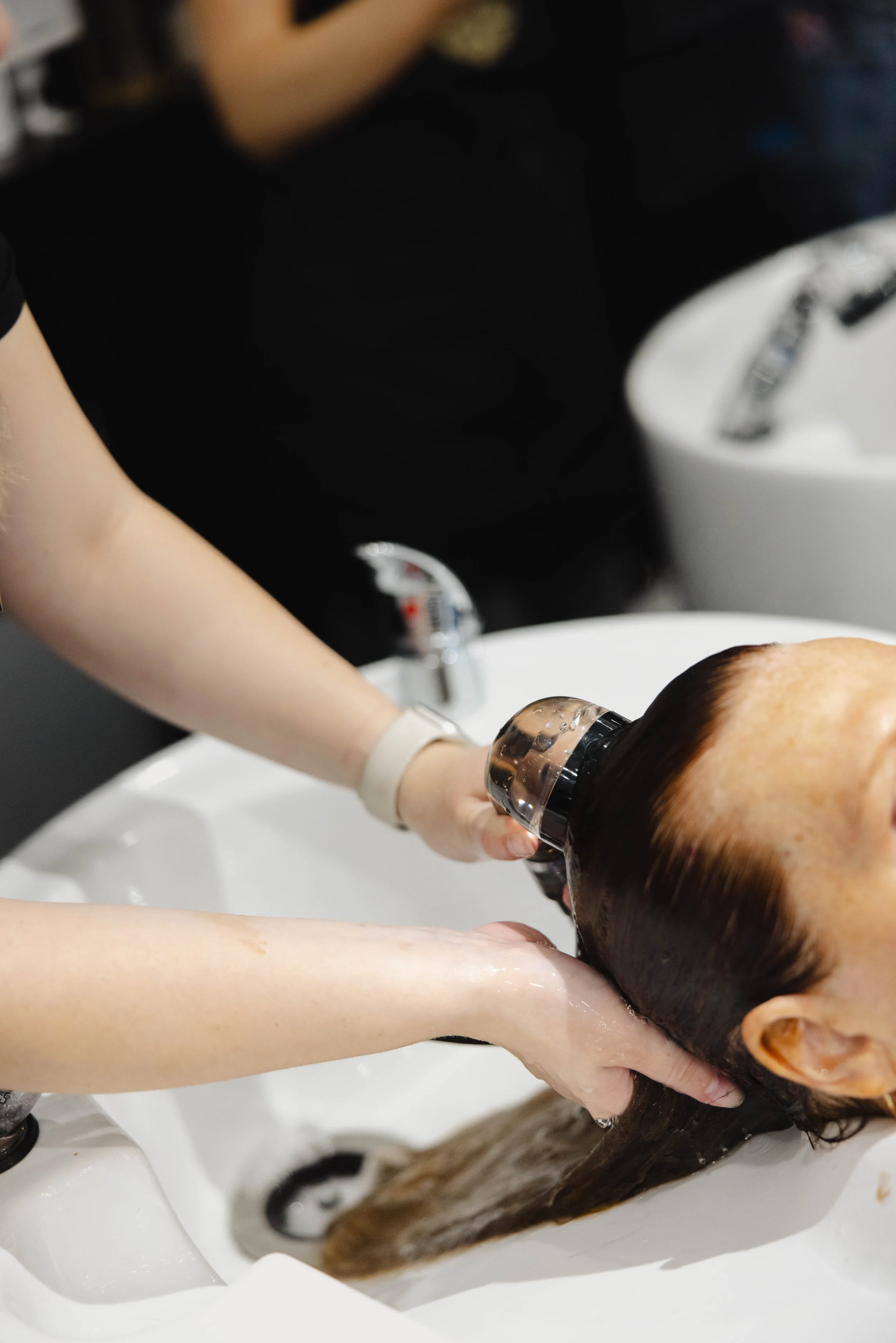 A person getting their hair washed at a salon sink while a stylist rinses their hair.