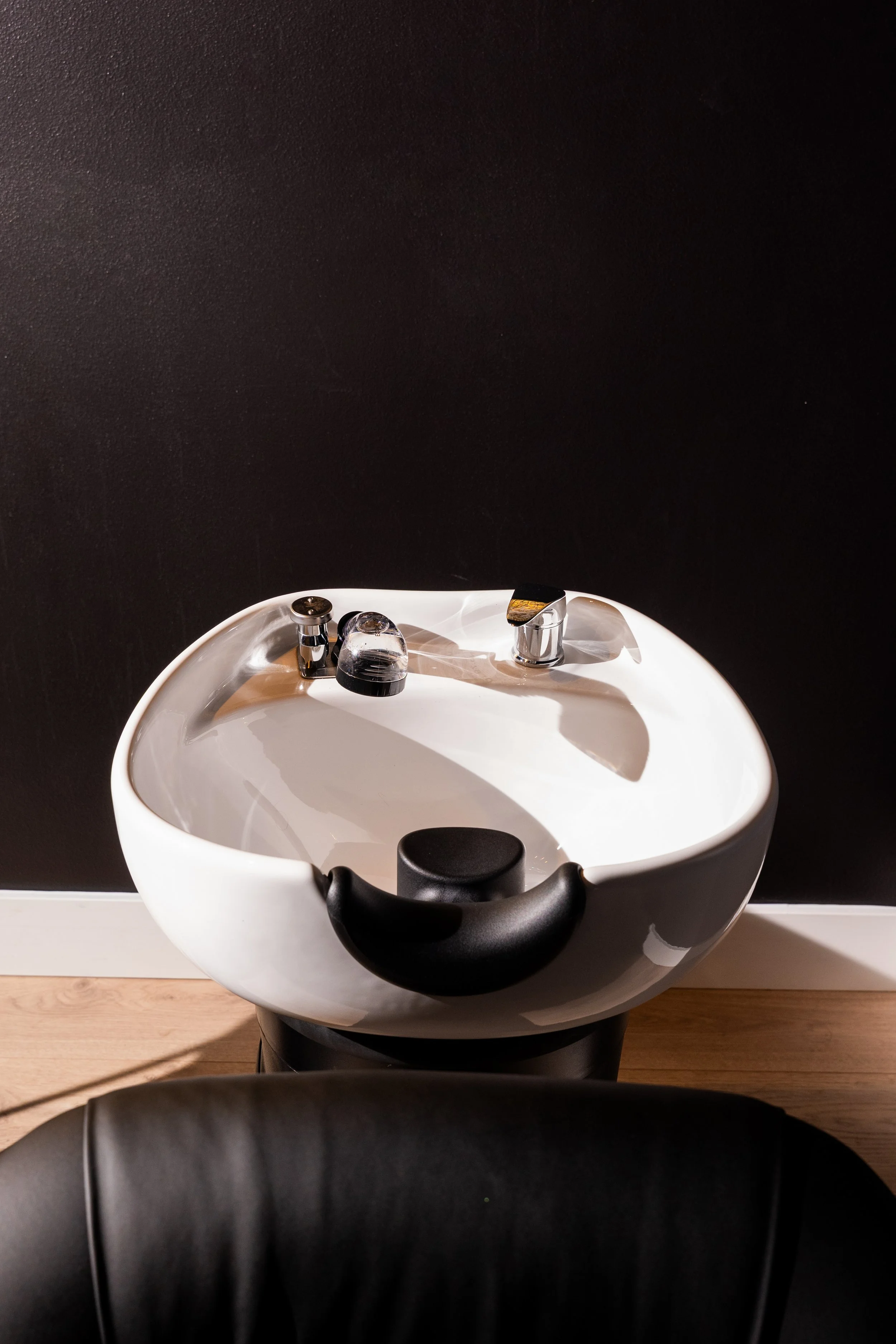 A white salon sink with a black cushioned headrest, situated in front of a black wall, with a chair in the foreground.