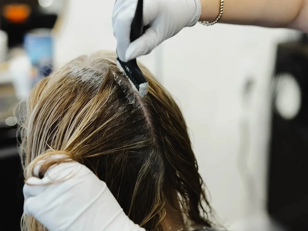 A hairdresser applies hair color to a woman's hair in a salon, wearing white gloves and a bracelet.