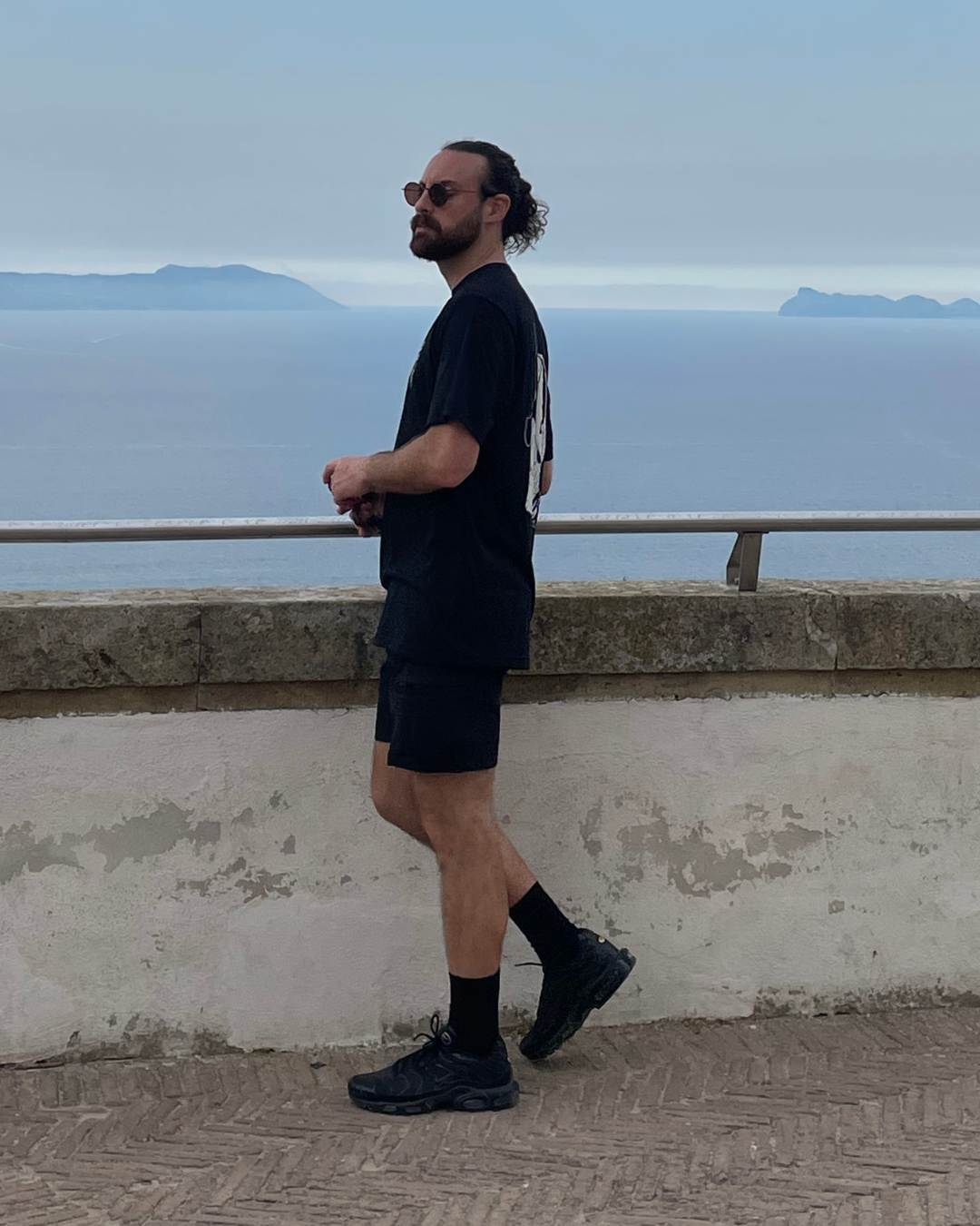 Man with curly hair, beard, sunglasses wearing black T-shirt, shorts, and sneakers standing on a seaside walkway, with ocean and islands in the background.