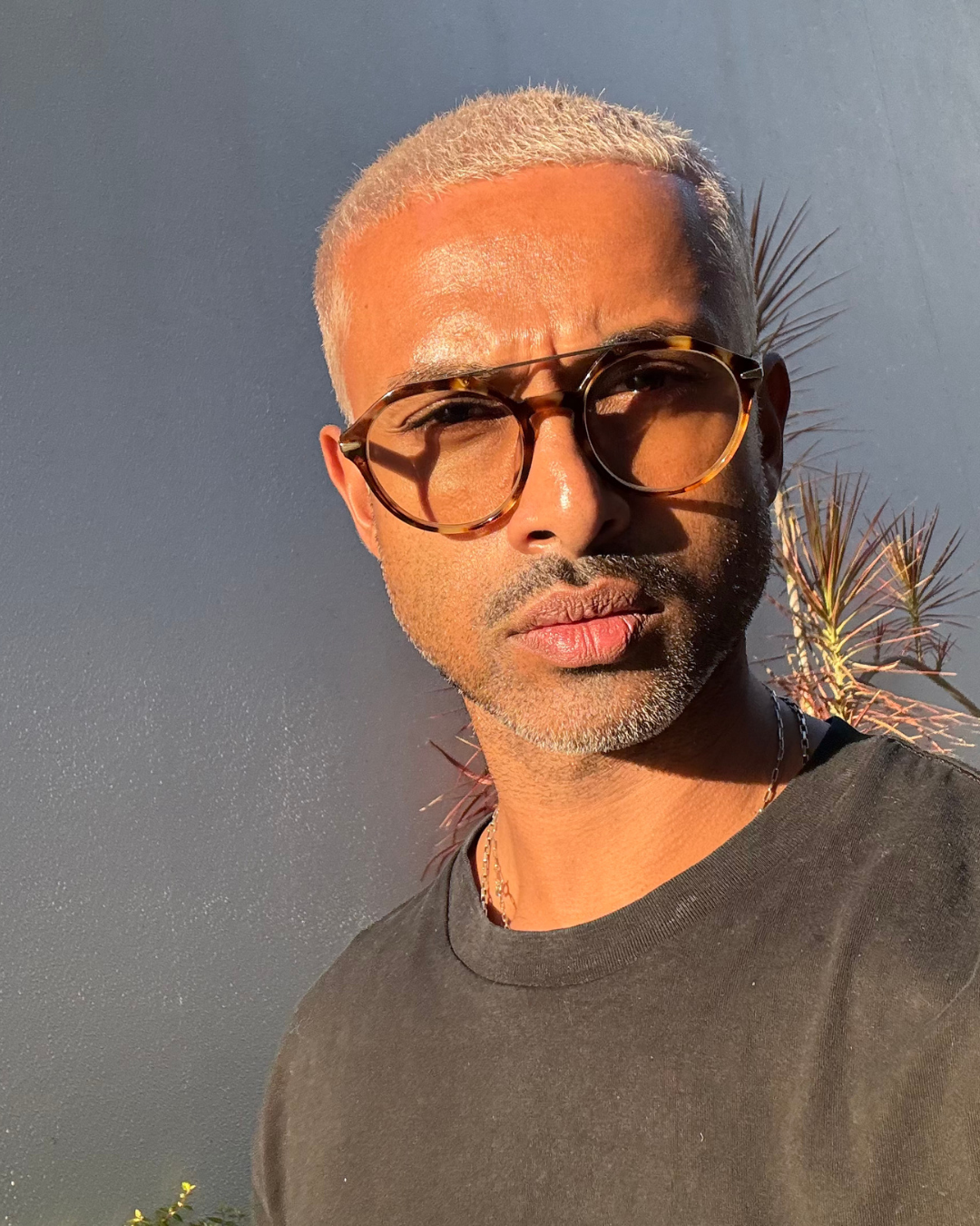 A man with short blond hair, wearing sunglasses and a black t-shirt, standing outdoors with a dark sky and palm leaves in the background.