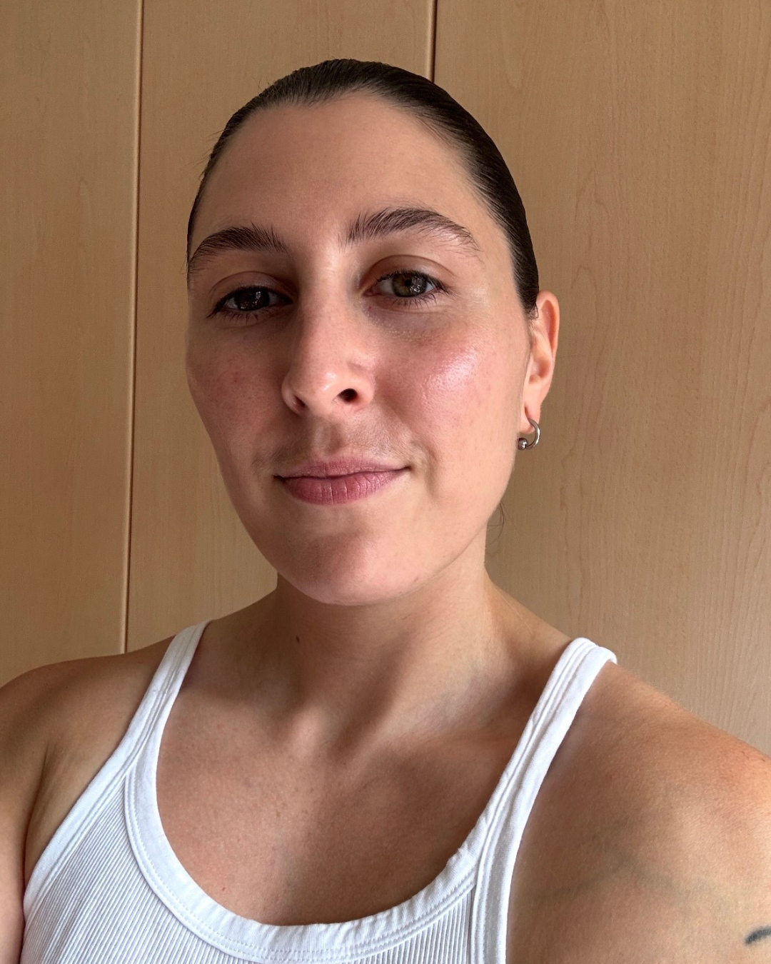 Close-up of a woman with dark hair pulled back, wearing a white tank top, standing in front of a wooden background.