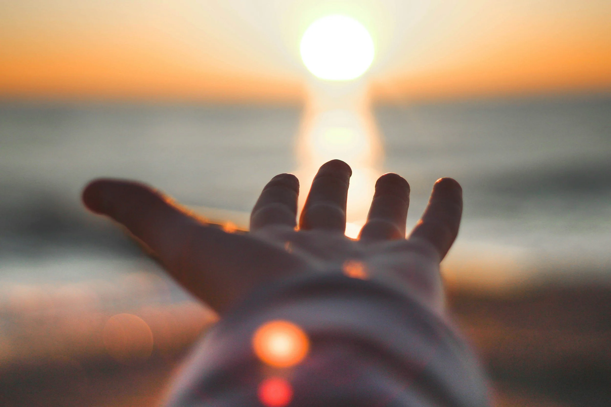 A person's hand reaching towards the setting sun in a gesture of hope, over the ocean at sunset.