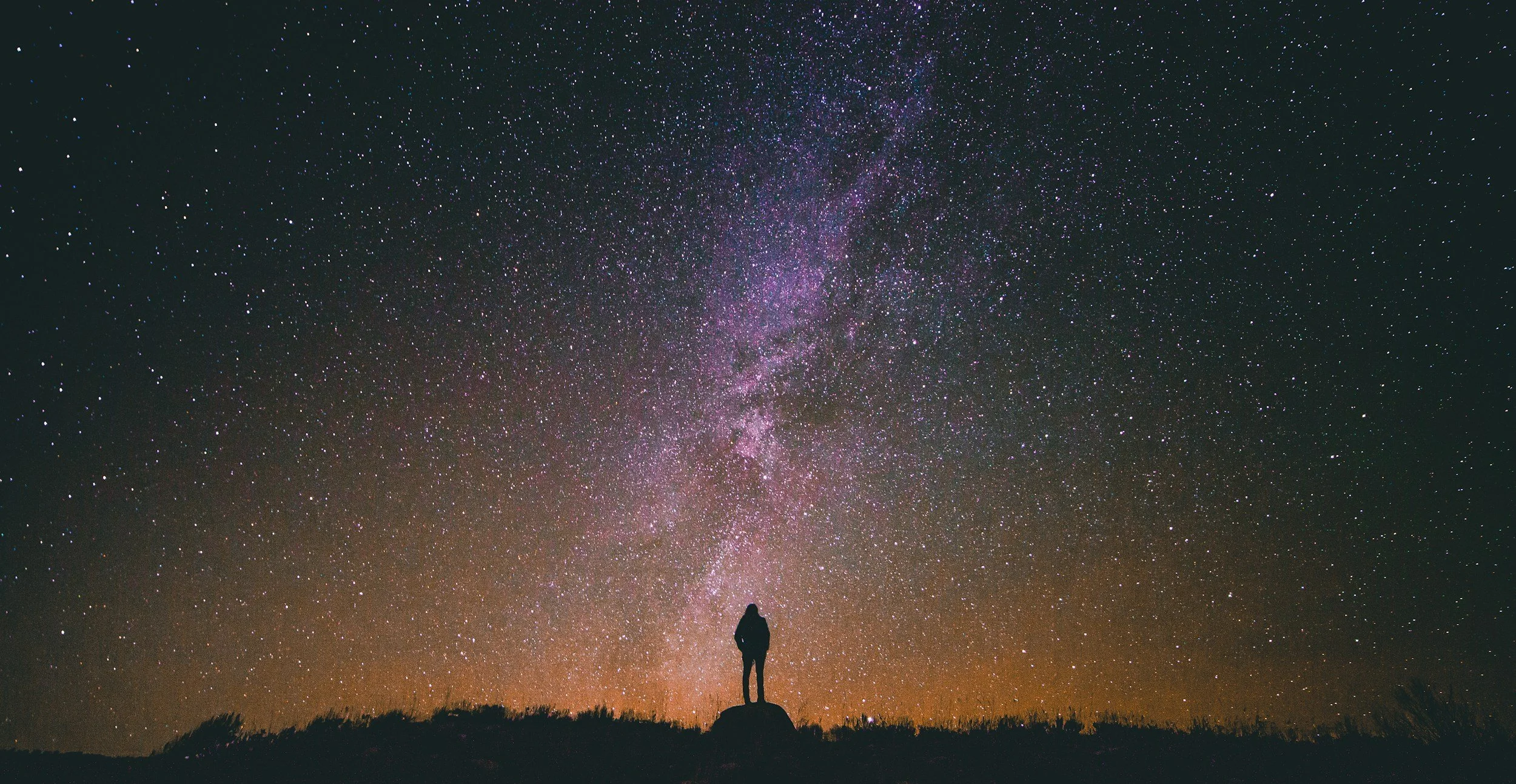 A person pondering their existence, standing on a hill staring upwards at a beautiful a starry night sky, with the Milky Way galaxy visible.