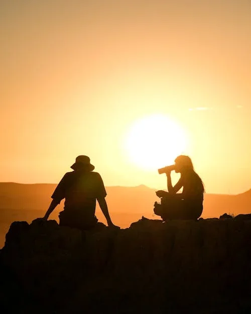 Two people sitting on a hill during sunset, one watching the other one with binoculars as if examining them.