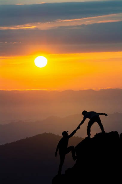 Two people silhouetted against a sunset, with one person extending their hand down to the other person below them to help the other climb up a mountain.