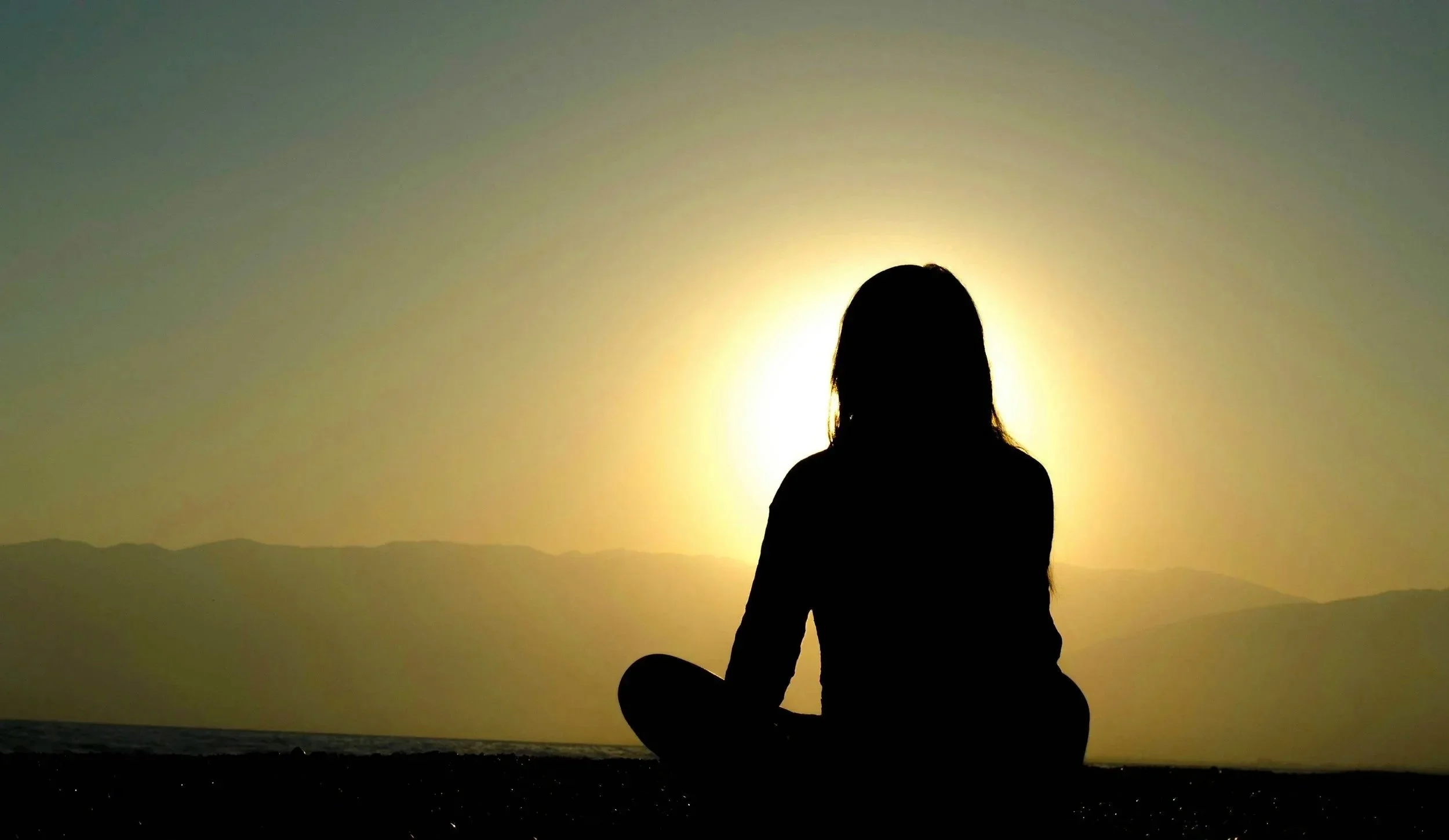 A peaceful silhouette of a person meditating watching a sunset with mountains in the background, evoking a sense of calm.