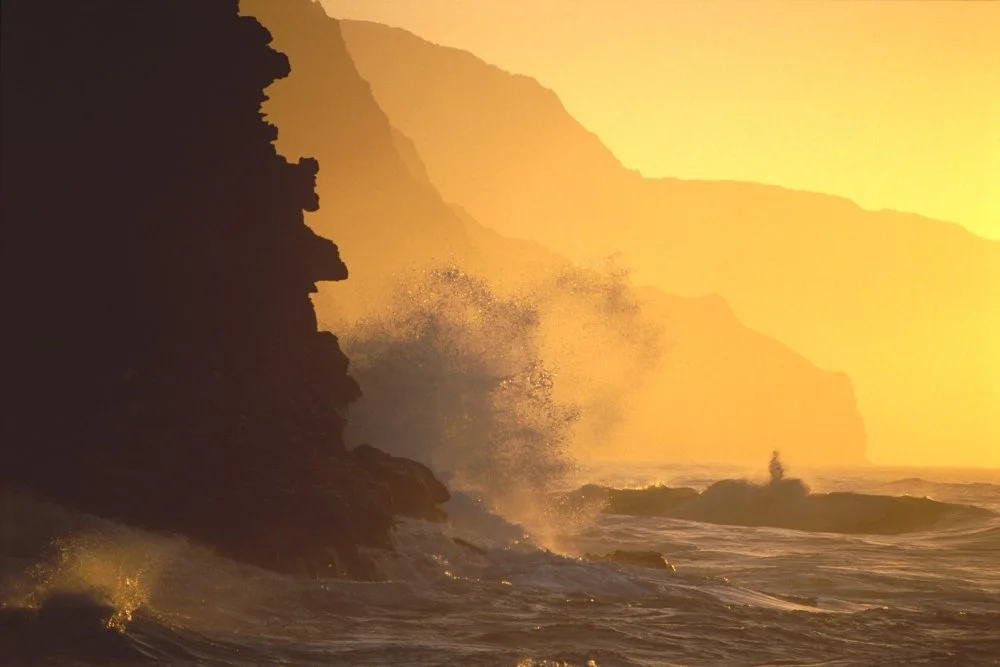 Sunset over rocky cliffs with ocean waves crashing against the shore.