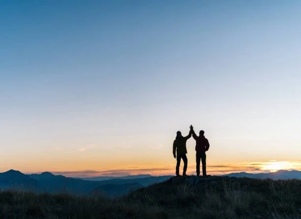 Two people standing on a hilltop at sunset, celebrating their climb together, silhouetted against a colorful sky, touching hands in a high-five.