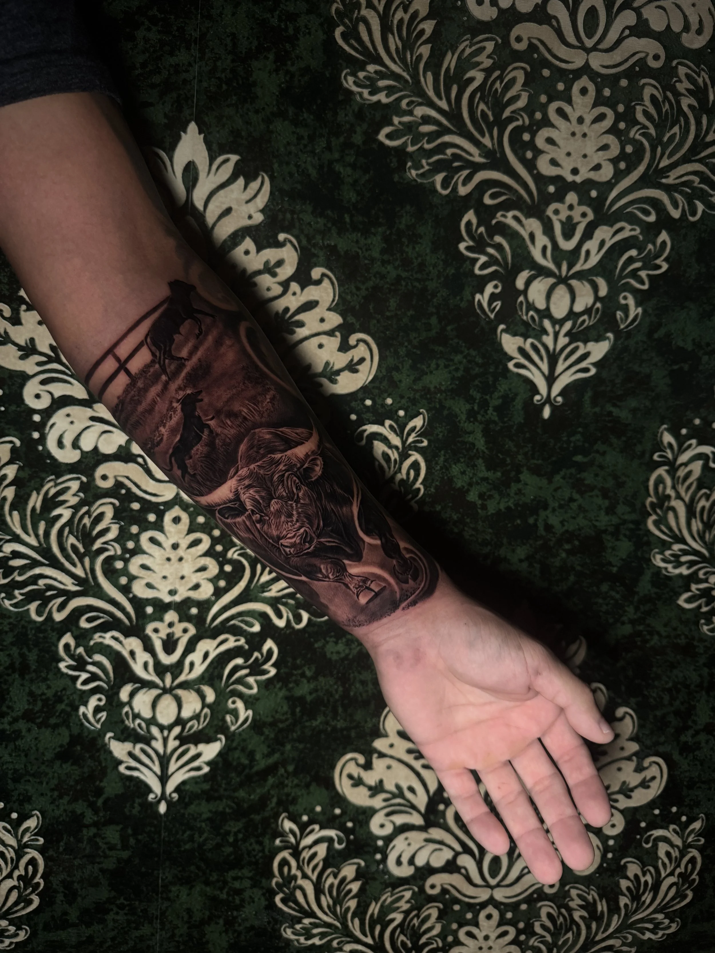 A person's arm with a detailed black and grey tattoo of a lion and a landscape, resting on a green and white ornate patterned surface.