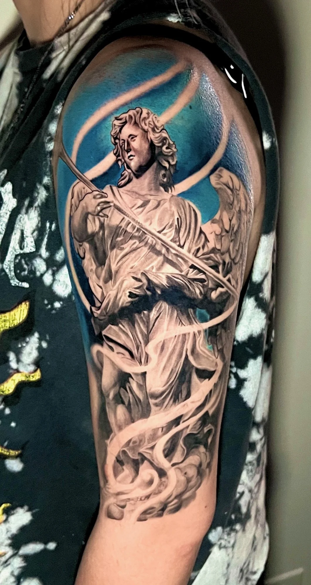 Tattoo of a grayscale angel holding a sword, with a blue and white swirling sky background, on a person's upper arm.