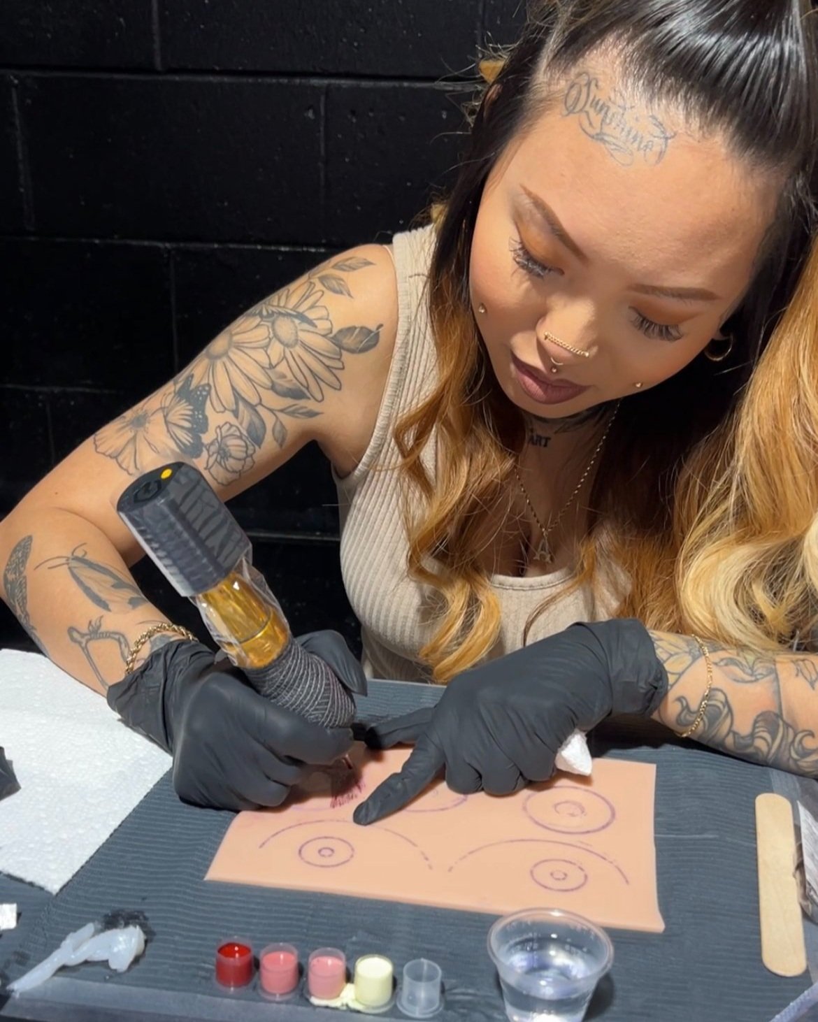 A woman with tattoos on her arms and face is tattooing a tattoo design on skin using a tattoo machine. She is wearing black gloves and has long, wavy hair.