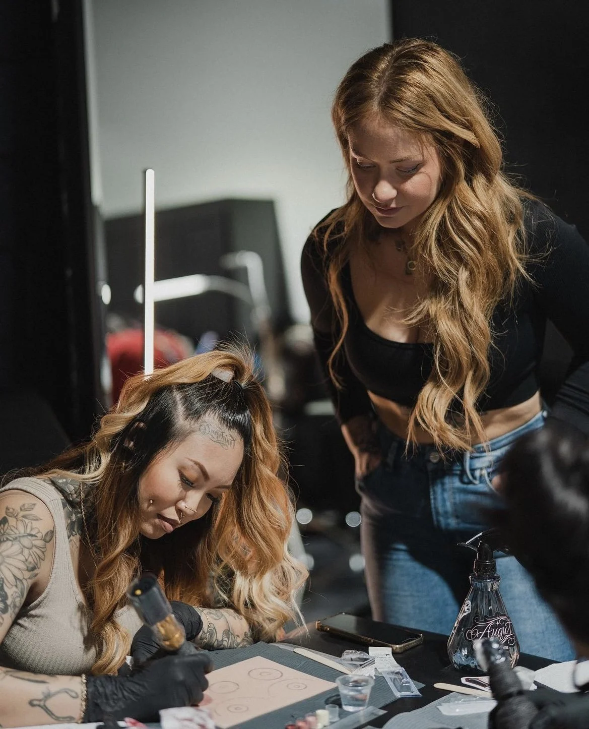 A woman with tattoos and long, wavy hair getting a tattoo on her arm while another woman with long, wavy hair and a black crop top looks on.