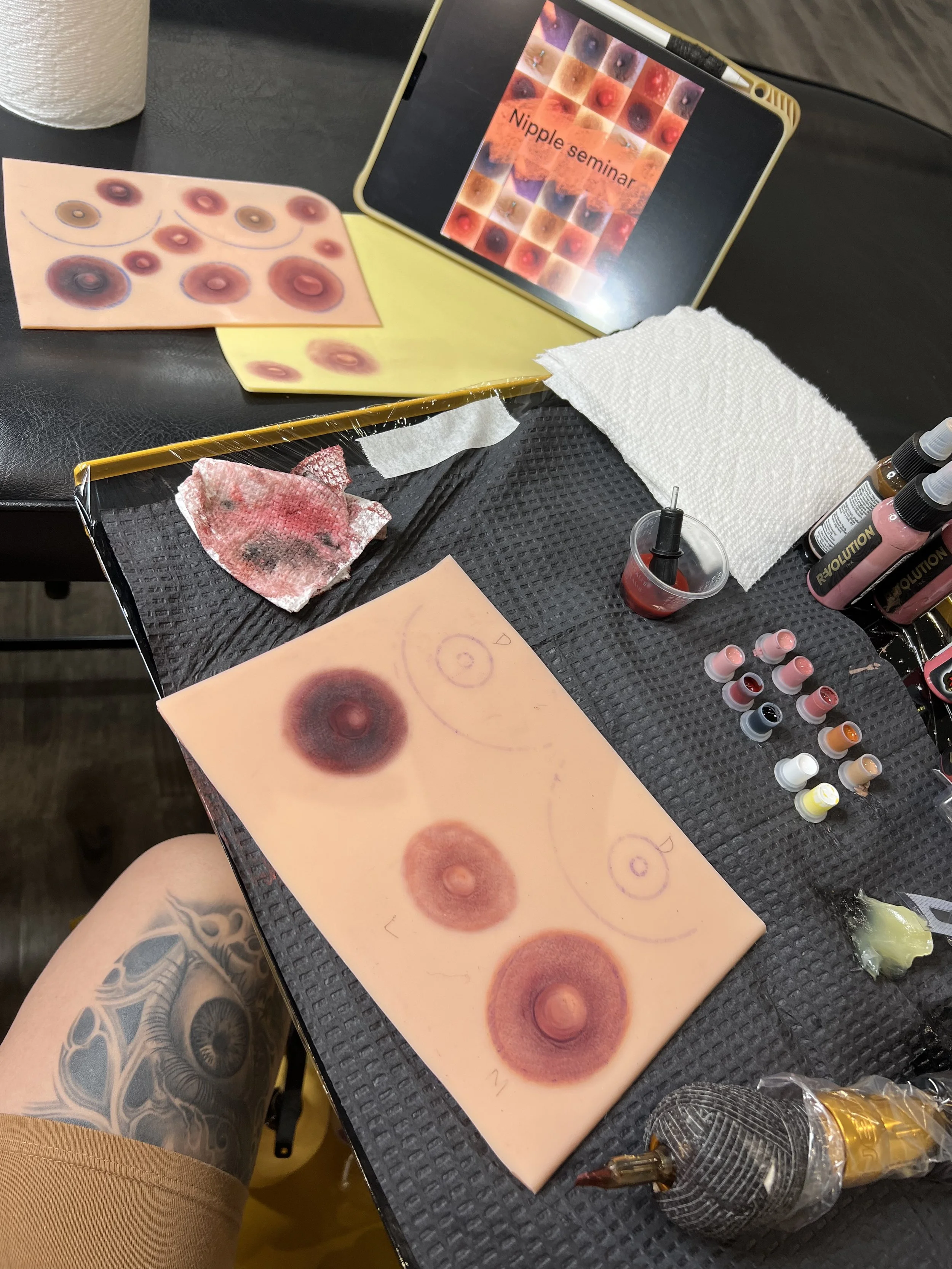 Tattoo practice sheet with multiple circles, ink bottles, paper towels, and a tablet displaying a diagram titled 'Nipple seminar' related to nipple tattooing.