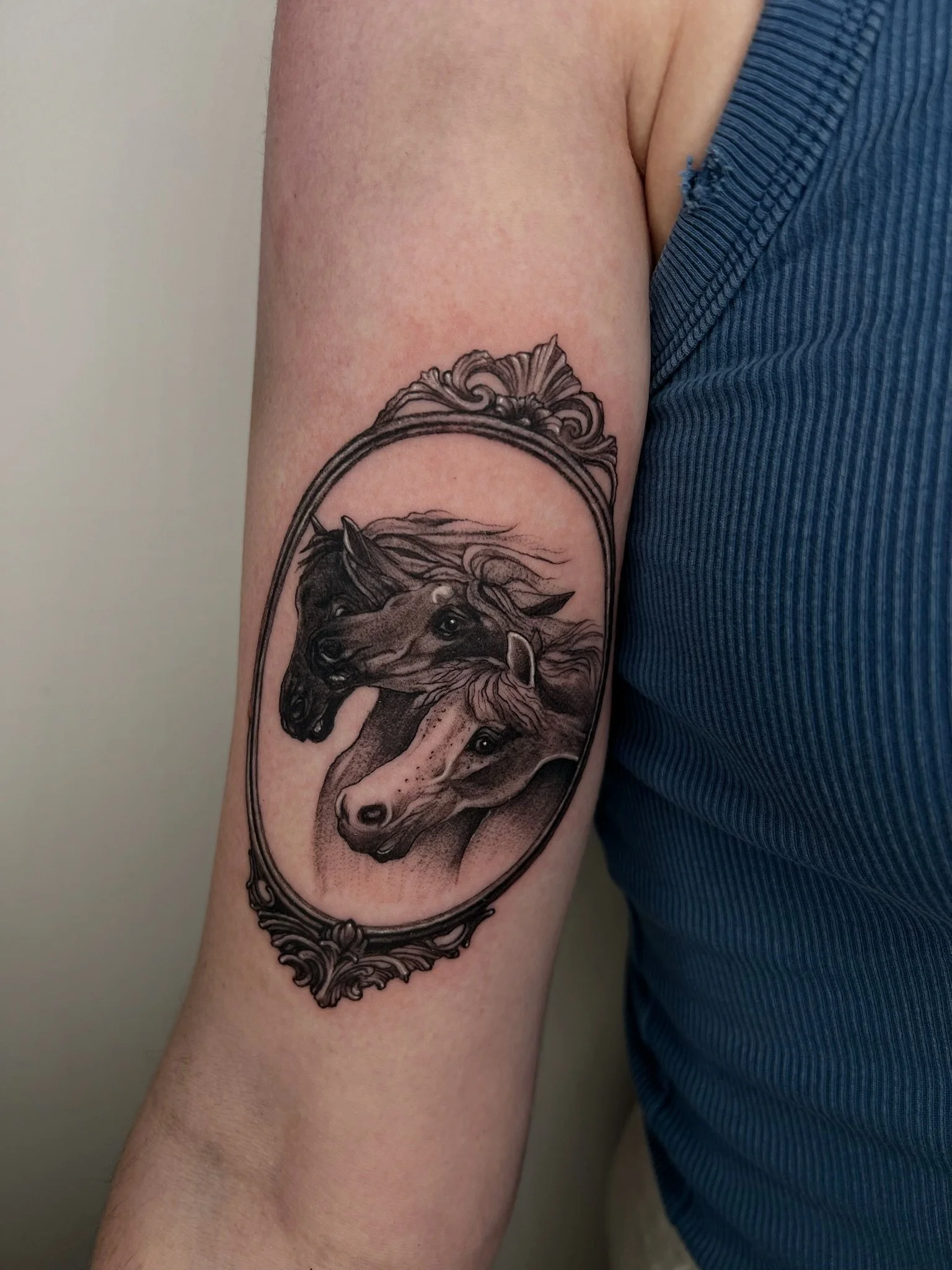 Tattoo of two horses' heads inside an ornate oval frame on person's upper arm.
