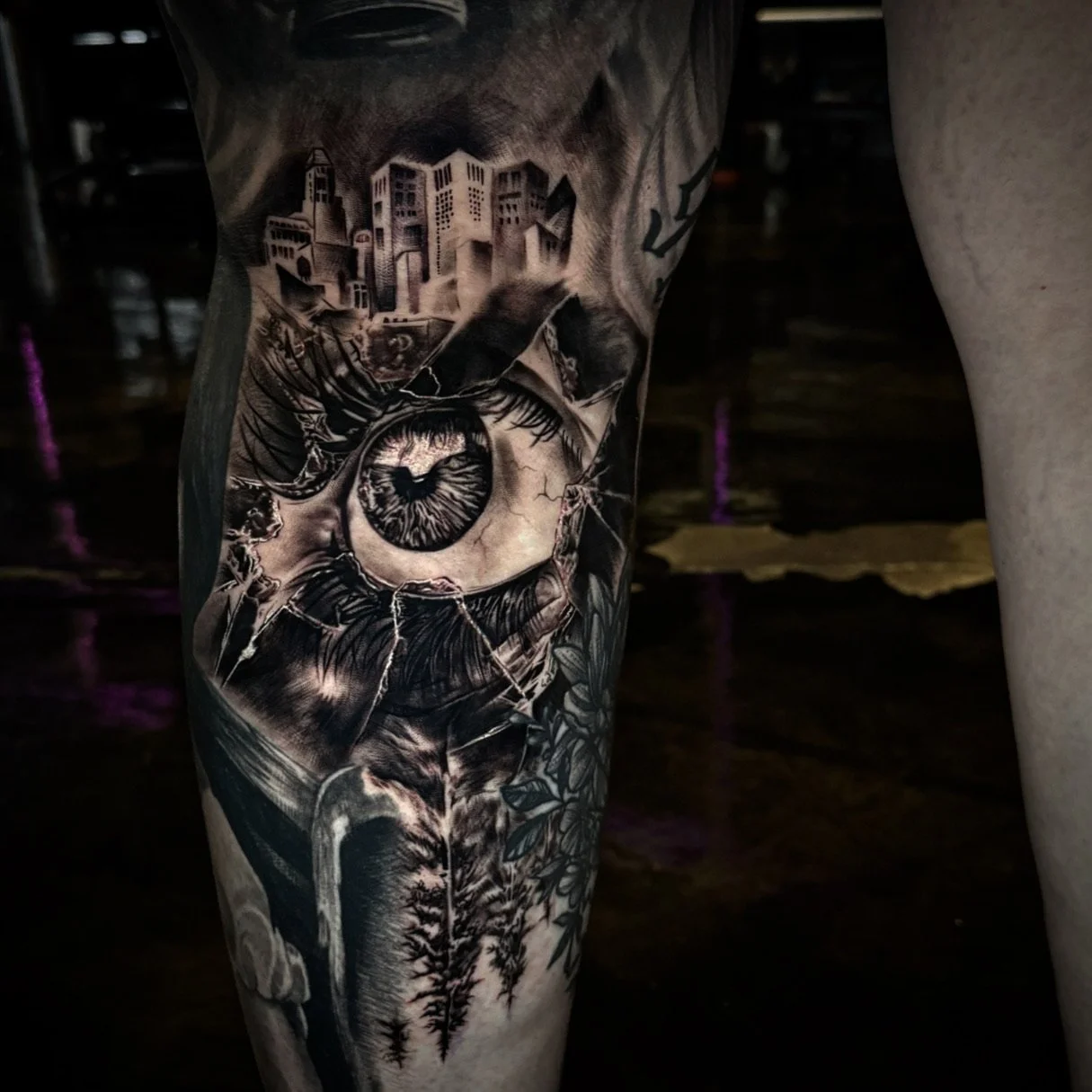 Close-up of a black and gray tattoo sleeve featuring a cracked eye, a cityscape, mountains, and trees.