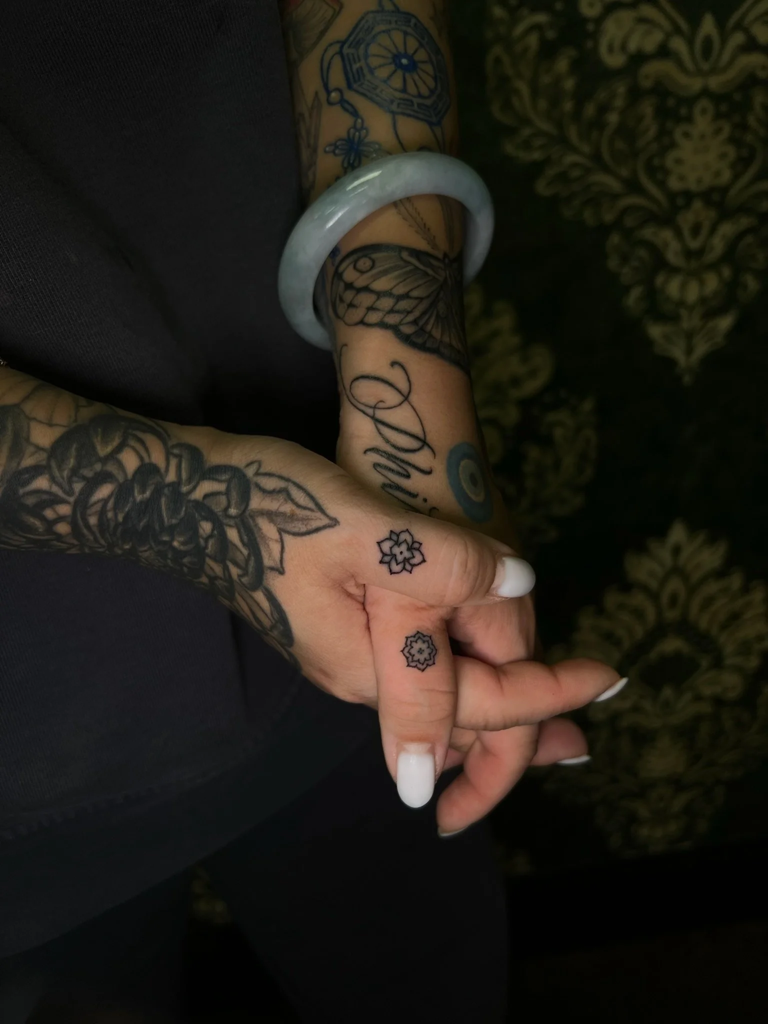 Close-up of a person's hand and forearm with tattoos, wearing a jade bangle. The tattoos include flowers, a butterfly, and various designs. The background features a dark, patterned fabric or wallpaper.